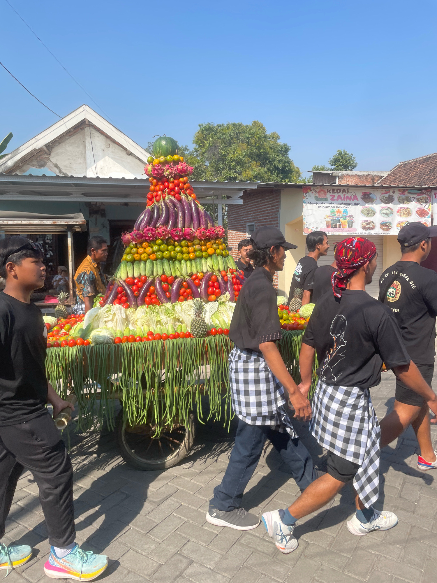 The image depicts a procession in the city of Kebon Waris, Indonesia. The main subject is a large, tiered display of fresh produce, including a watermelon at the top, followed by layers of dragon fruit, tomatoes, eggplants, oranges, cucumbers, cabbage, long beans, and pineapples. This elaborate arrangement is mounted on a wheeled cart, decorated with hanging long beans. Several young men, dressed in black t-shirts and plaid sarongs or wraps, are walking alongside the cart, holding hands in a synchronized manner. One individual is wearing a red and black patterned headwrap. In the background, there are buildings with tiled roofs, a banner with Indonesian text advertising "KEDAI ZAINAB" (ZAINAB'S SHOP) and images of food, and trees. The scene is set under a clear blue sky on a paved surface, suggesting a daytime outdoor event.