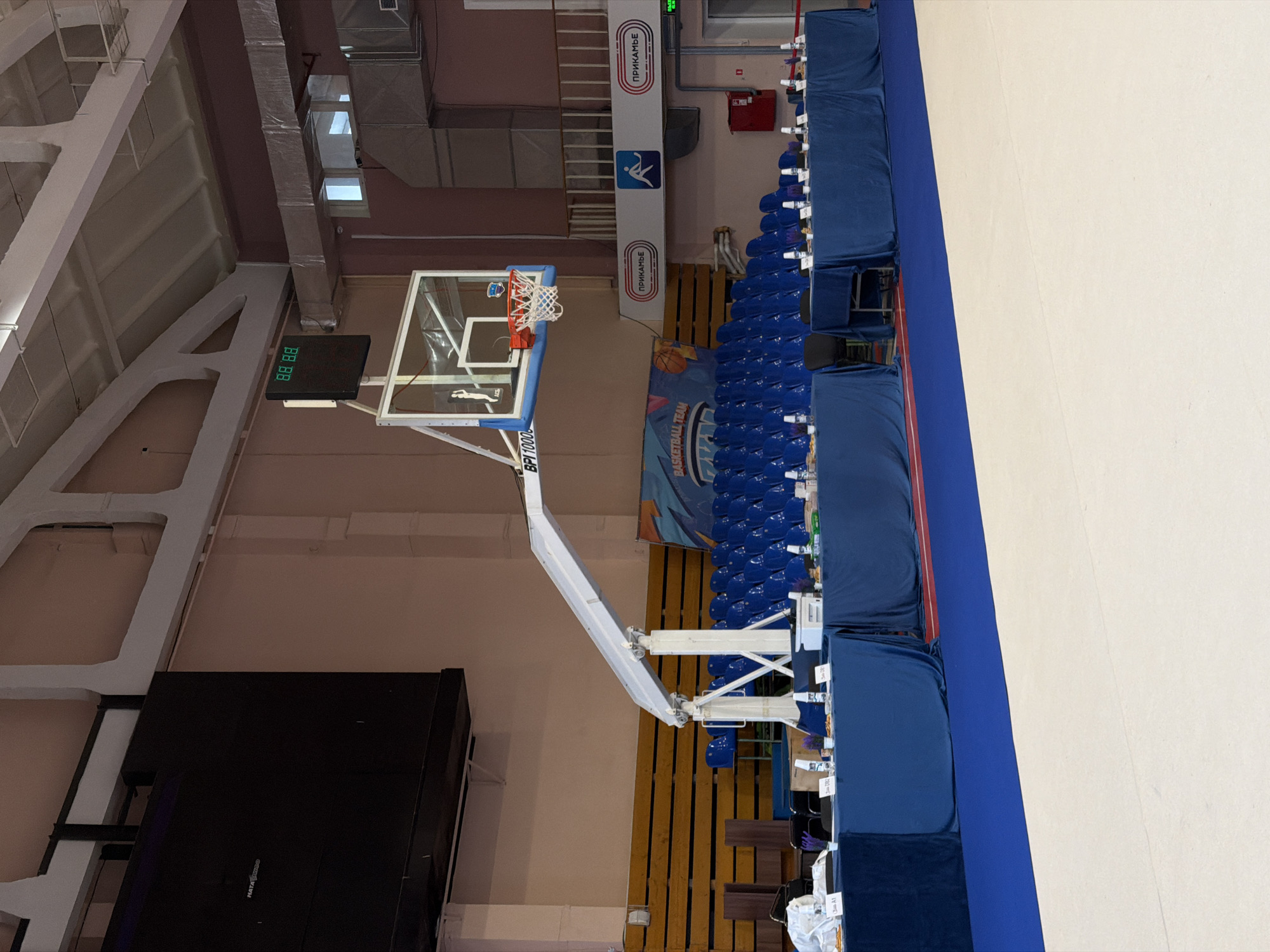 An indoor basketball court in Perm, Russia features a basketball hoop with an electronic scoreboard. Blue seating is visible next to the wall. Wall banners display the word "ПРИКАМЬЕ".