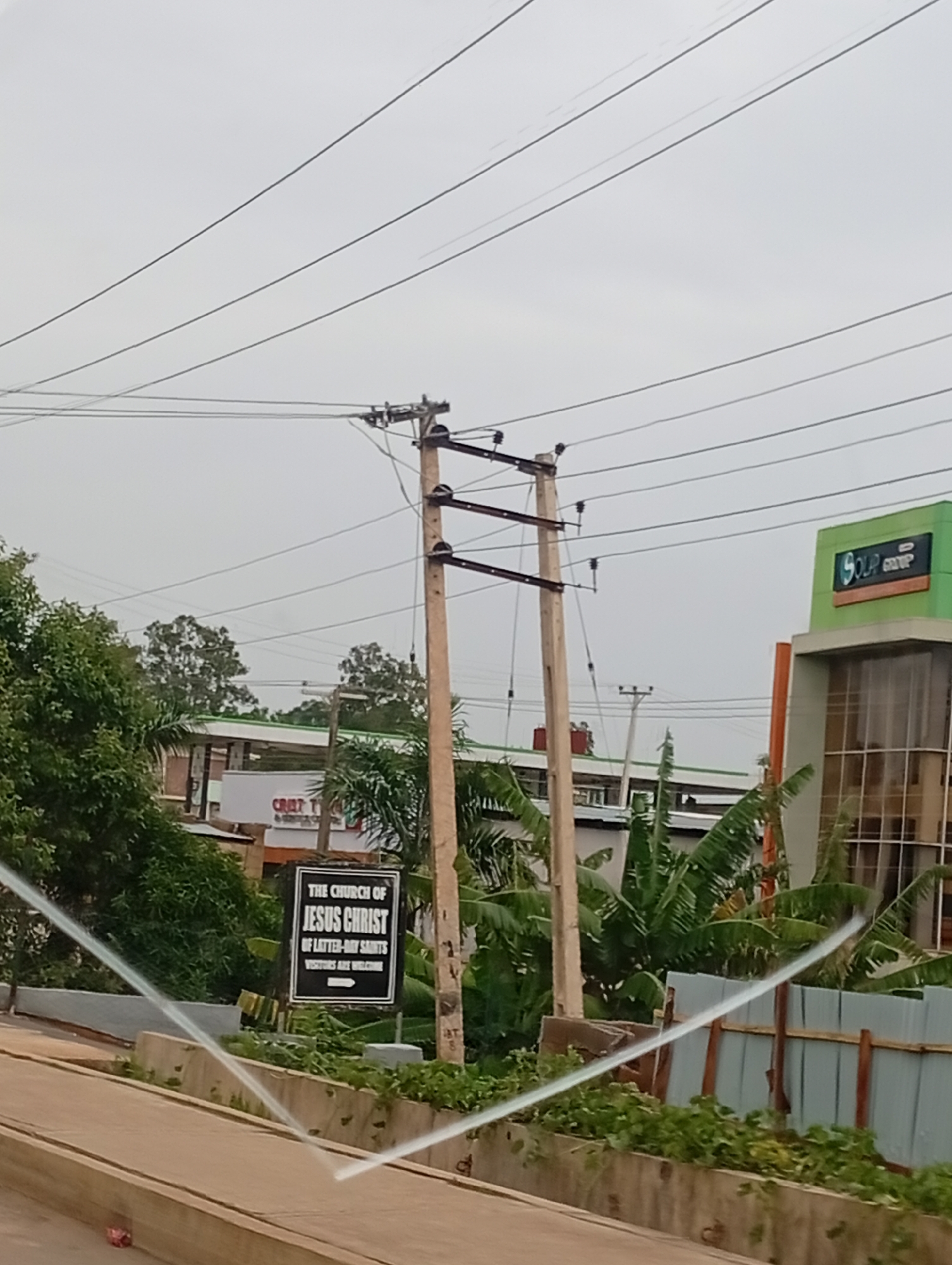 The image captures a street scene in Jos, Nigeria, with a prominent sign for "THE CHURCH OF JESUS CHRIST OF LATTER-DAY SAINTS" on the left. The sign is black with white text and an arrow pointing to the right. Behind the sign, lush green trees and banana plants are visible. In the background, a multi-story building with a green facade and the logo "SOLAR GROUP" can be seen on the right. Power lines crisscross the sky, supported by wooden utility poles. The foreground shows a concrete barrier and a paved surface, possibly a road or sidewalk. The sky is overcast and appears to be daytime. There are no people or animals visible in the image, and no specific actions or interactions are depicted.