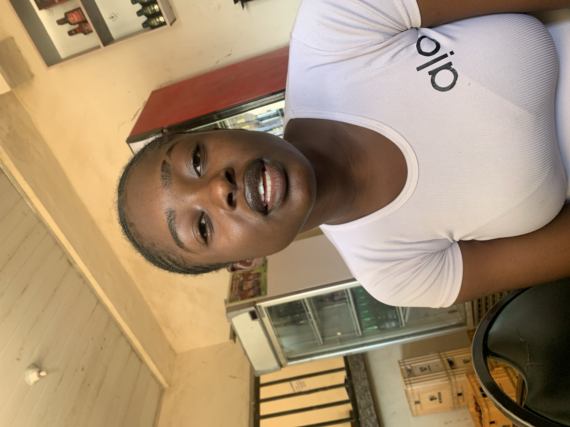 A young Black woman with dark skin and braided hair is positioned in the center of the frame, looking directly at the camera. She is wearing a white ribbed t-shirt with the black logo "alo" on the right side of her chest. Her lips are slightly parted, revealing her teeth, and she appears to be speaking. The background is dimly lit and suggests an indoor setting, possibly a shop or restaurant in New Stadium, Nigeria. To her left, a glass-fronted refrigerator is visible, stocked with bottles. In the background to the right, a red refrigerator stands next to a shelf displaying bottles. The ceiling is made of wooden planks, and a light fixture hangs from it. The overall impression is that of a casual, candid shot taken in a local establishment.