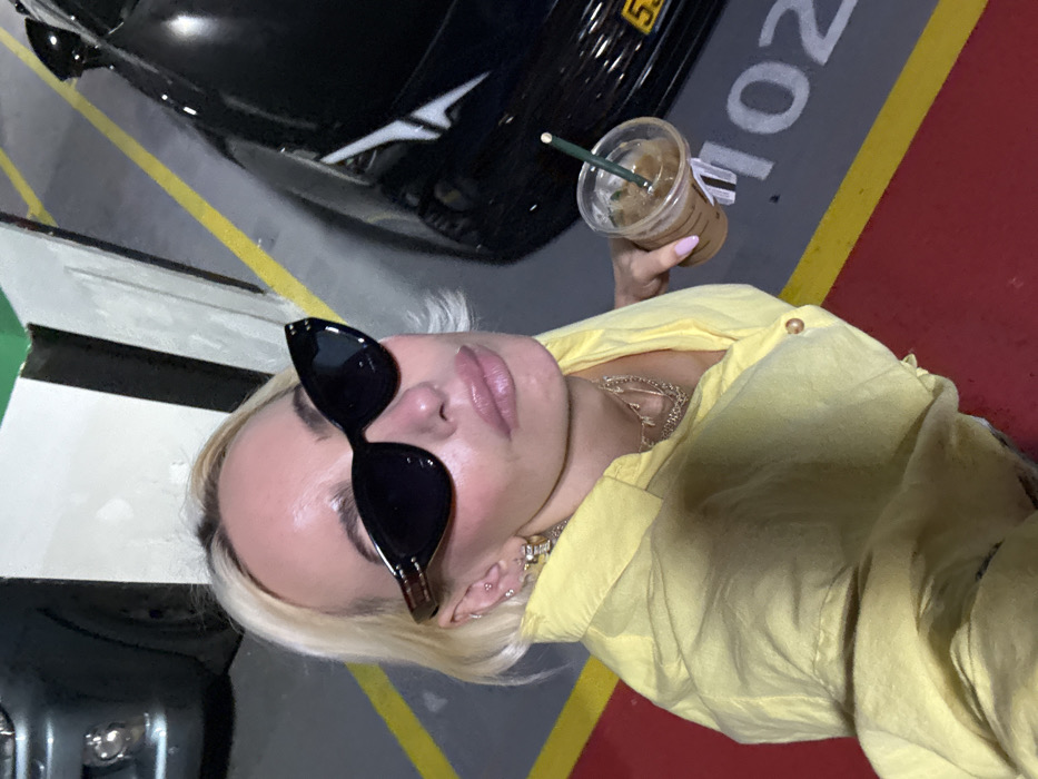 The image depicts a fair-skinned individual with short, bleached blonde hair and visible dark roots, captured in a high-angle selfie in an indoor parking garage. The person is wearing dark cat-eye sunglasses, a light yellow collared shirt, multiple ear piercings with various earrings, and layered gold-colored necklaces. In their right hand, they hold a clear plastic cup containing an iced coffee with a green straw. The visible hand has manicured nails with light pink polish.
The environment is a parking garage, characterized by a grey asphalt or concrete floor with yellow painted demarcation lines, a prominent red painted section in the immediate foreground, and white painted numbers "102" indicating a parking spot. A partially visible black luxury vehicle with a yellow license plate displaying "53" is parked to the individual's right. To the far left, part of a dark grey vehicle's front bumper is visible. The background features concrete walls with sections painted white and green. The location is an unspecified parking facility within the city of Dubai, United Arab Emirates.