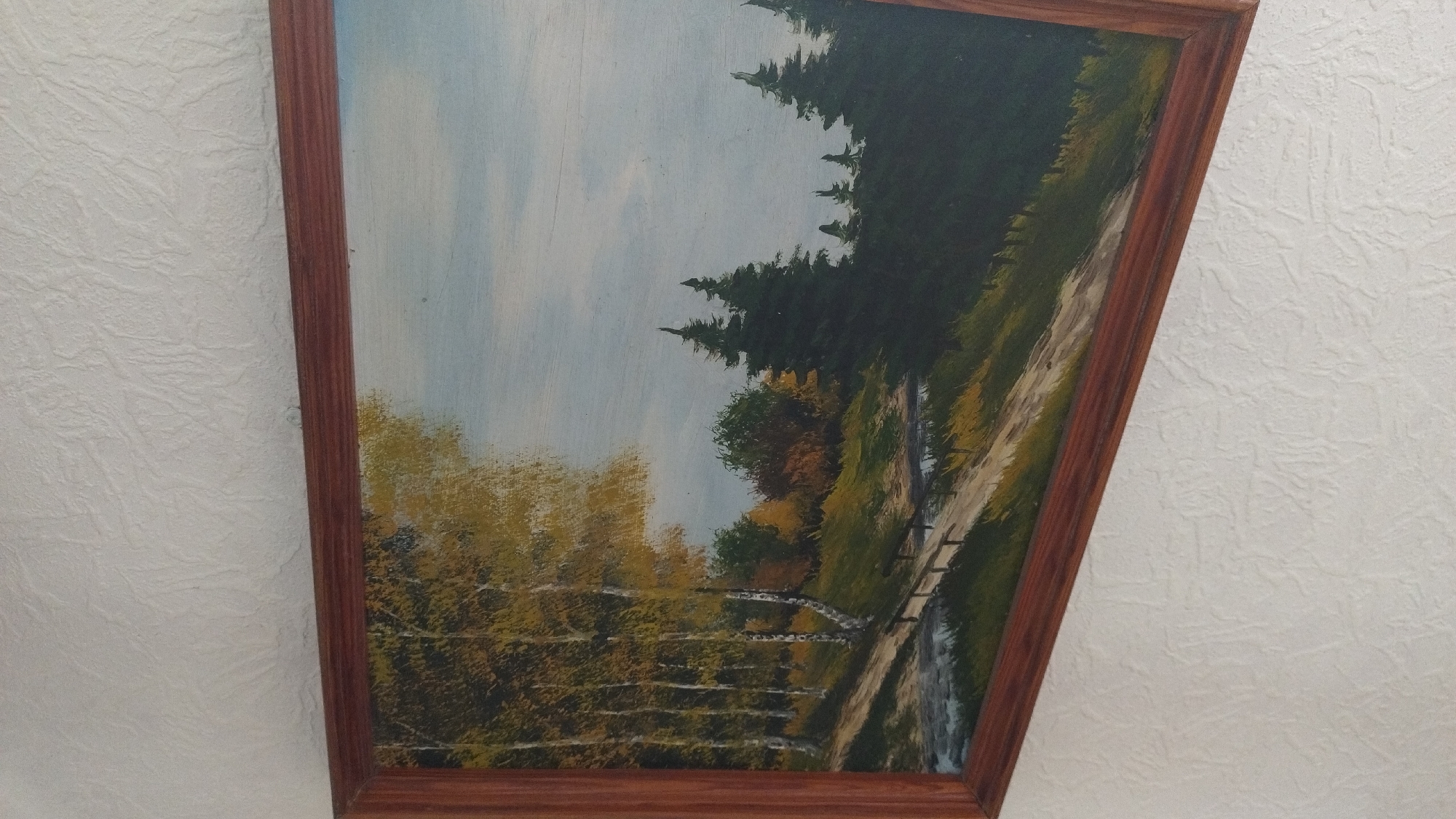 A framed oil painting depicts an autumn landscape scene with a dirt path winding through a grove of birch trees with yellowing foliage on the left. The path leads to a small body of water. In the middle ground, there are more trees with autumnal colors, and on the right, a line of dark green evergreen trees. The sky is pale blue with wispy clouds. The painting is hung on a textured white wall.