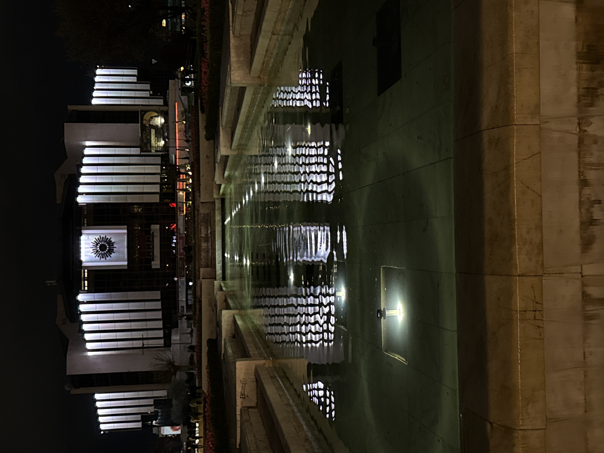 The image shows a modern building with horizontal illuminated stripes reflected in a long pool of water at night. The building has a circular emblem on one of its sections. The pool is lined with stone or concrete structures, and there are recessed lights on the side of the pool. The building appears to be situated in an urban area, as suggested by the surrounding structures and lighting. The location is Unknown, Unknown.