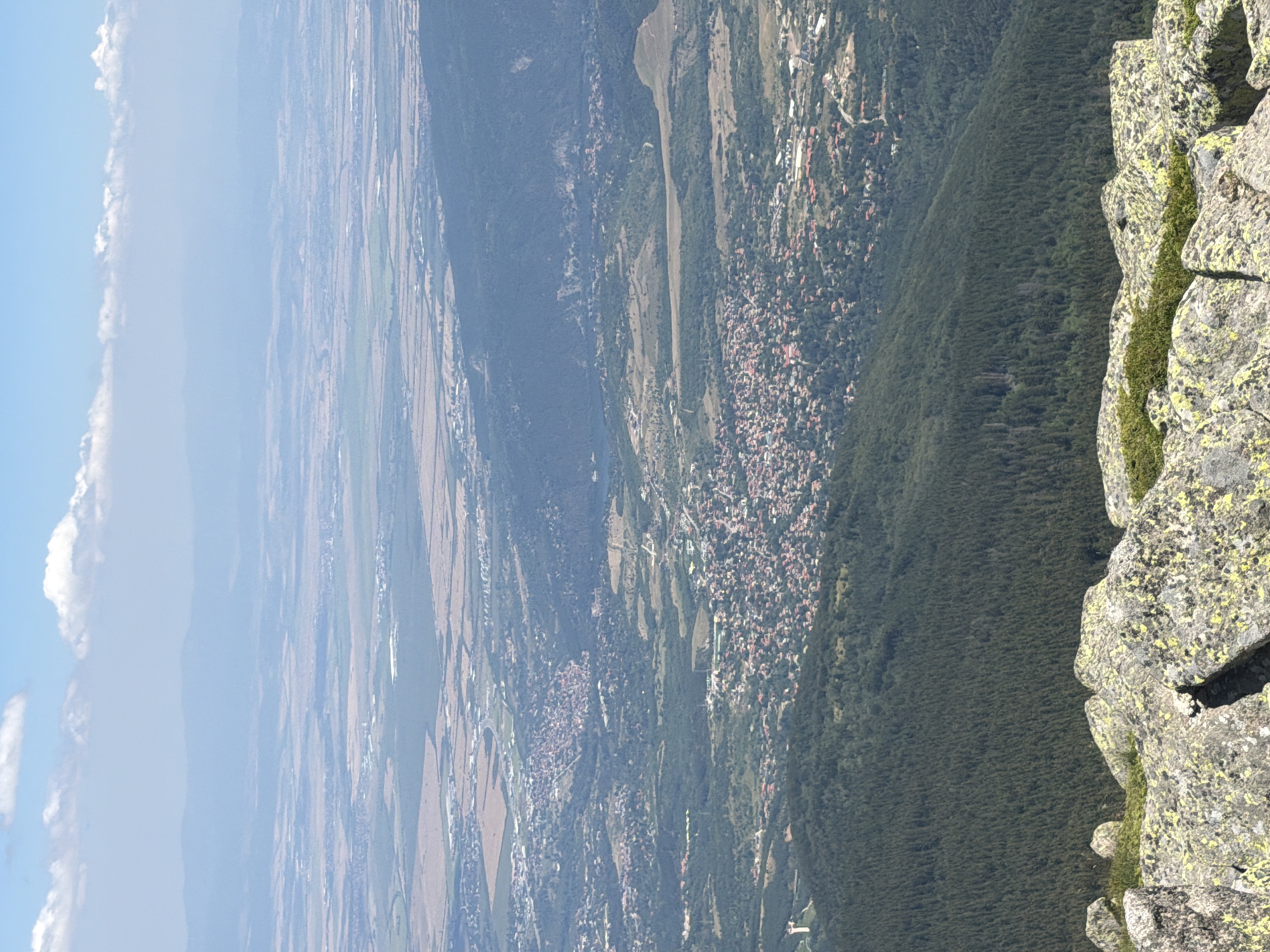 The image is a high-angle, wide shot of a landscape with a city in the middle ground. In the foreground, large, lichen-covered rocks and sparse green vegetation are visible. The middle ground is dominated by a densely populated town with red-tiled roofs, nestled within a valley and surrounded by lush green forests. This town appears to be Bistritsa, Bulgaria, given its geographical setting. Beyond the town, the landscape opens up to a vast plain dotted with agricultural fields and smaller settlements, stretching towards distant, hazy mountains under a bright blue sky with scattered white clouds. The overall impression is one of a sprawling, populated area within a natural, mountainous environment.