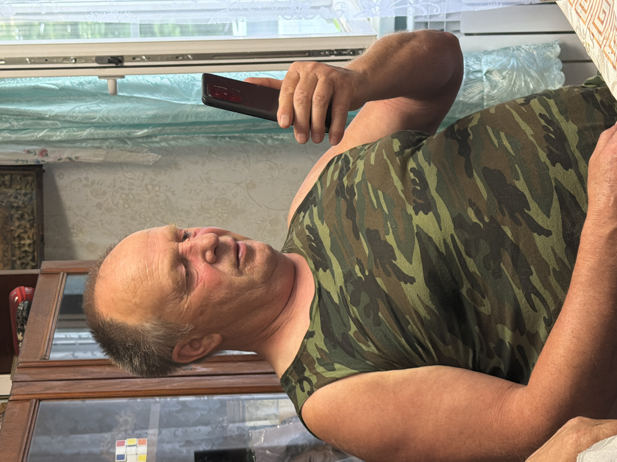 A man in a camouflage tank top is holding a smartphone in his right hand. The phone is black and has "Redmi" written on its back. The man's face is turned upwards and to the right, with his eyes closed. He appears to be relaxed or asleep. The background shows a window with light-colored curtains and a wooden piece of furniture with glass doors. The image is taken indoors, likely during the day, given the natural light from the window. The setting suggests a home or a casual indoor environment. The man's skin is flushed, and he has wrinkles on his face, indicating he is an older adult. The overall mood is peaceful and still.