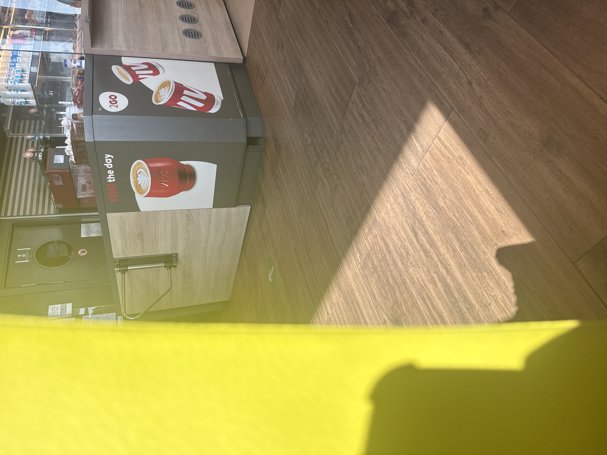 The image depicts a service counter area, likely within a cafe or convenience store. The counter has a modern design with wood-grain paneling and dark grey accents. On the counter, there are graphics of coffee cups and branding that reads "VIVA the day" and "2GO". Behind the counter, shelves are visible stocked with various bottles, and a red coffee machine is present. To the left of the counter, a bright yellow object, possibly a wall or partition, occupies a significant portion of the frame, casting a shadow on the wooden floor. The floor is made of wood-look tiles laid diagonally. A metal turnstile and a door with a circular window are also visible in the background. The lighting suggests it is daytime, with sunlight creating strong shadows across the floor. There are no people visible in the image. The location is stated to be in Drazhevo, Bulgaria.
