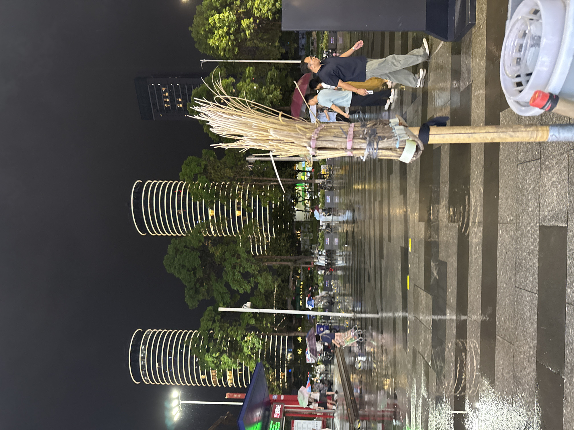 This image depicts a street scene at night in Tianhe District, China, likely after a rainfall, as indicated by the wet pavement and reflections. In the foreground, a bundle of dried reeds, tied together with pink and dark blue bands and attached to a wooden pole, stands vertically. To the right of the reeds, a white plastic mop bucket with a red and black handle is partially visible.
In the mid-ground, several people are walking, some under umbrellas. A man in a dark blue polo shirt and khaki pants is walking towards the right, looking forward. To his left, a woman in a light blue t-shirt and yellow pants is walking with her head down, holding an umbrella. Further back, other individuals and groups can be seen navigating the wet street, with some pushing carts.
The background features two distinctive cylindrical buildings with white horizontal light strips illuminating their facades, creating a modern architectural aesthetic against the dark night sky. Lush green trees are interspersed between and around the buildings, adding a natural element to the urban landscape. A tall, dark building with illuminated windows is also visible in the far background. Streetlights and architectural lighting contribute to the overall illumination of the scene.