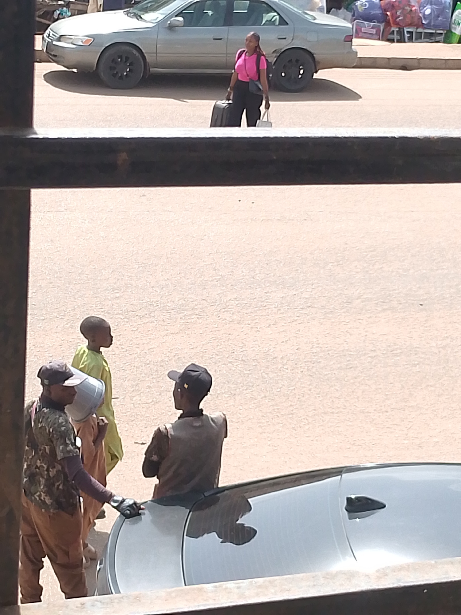 A woman in a pink t-shirt and black pants walks with luggage beside a silver sedan. The scene appears to be outdoors, possibly on a street or in a parking lot, with a dusty, paved surface. In the foreground, three people are gathered near a black car. Two of them are wearing caps, and one is holding a large, light-colored bucket. The person in the center of this group is wearing a bright yellow shirt and appears to be a child. The overall lighting suggests it is daytime, likely with clear weather. There is no visible text in the image. The setting, with the cars and the casual arrangement of people, suggests an everyday street scene in Bukuru, Nigeria.