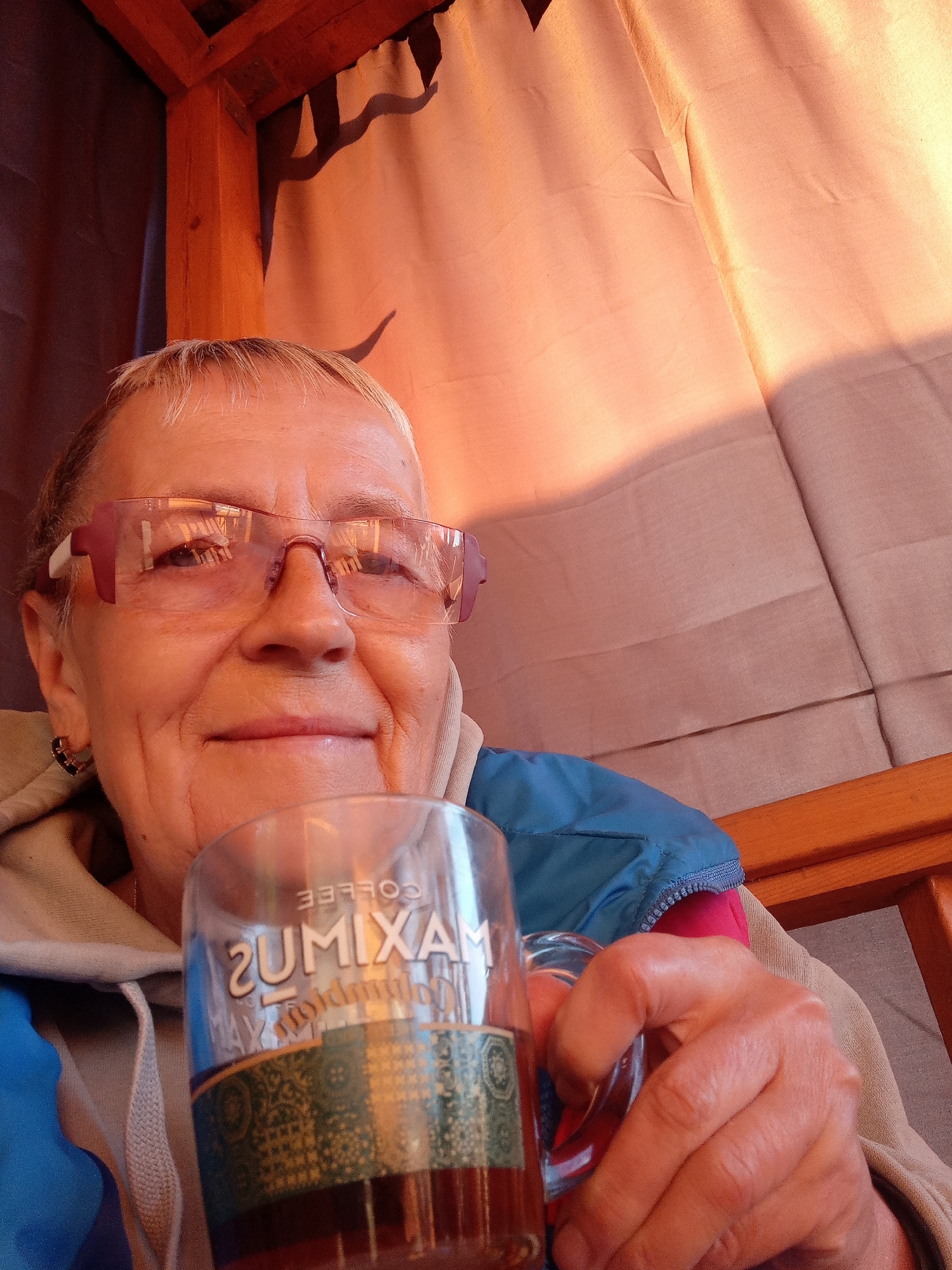 A middle-aged Caucasian woman with short blonde hair and pink-rimmed glasses is holding a glass of dark liquid. She is wearing a light-colored hooded jacket with blue and pink accents and a beige hoodie underneath. The woman is seated in an outdoor setting, possibly a gazebo, with wooden beams visible above and a light-colored draped fabric in the background. The lighting suggests it is either early morning or late afternoon, with warm, golden light casting shadows. The glass has text that reads "MAXIMUS COFFEE" in reverse. The woman is smiling slightly as she looks towards the camera. The scene appears to be a casual, relaxed moment.