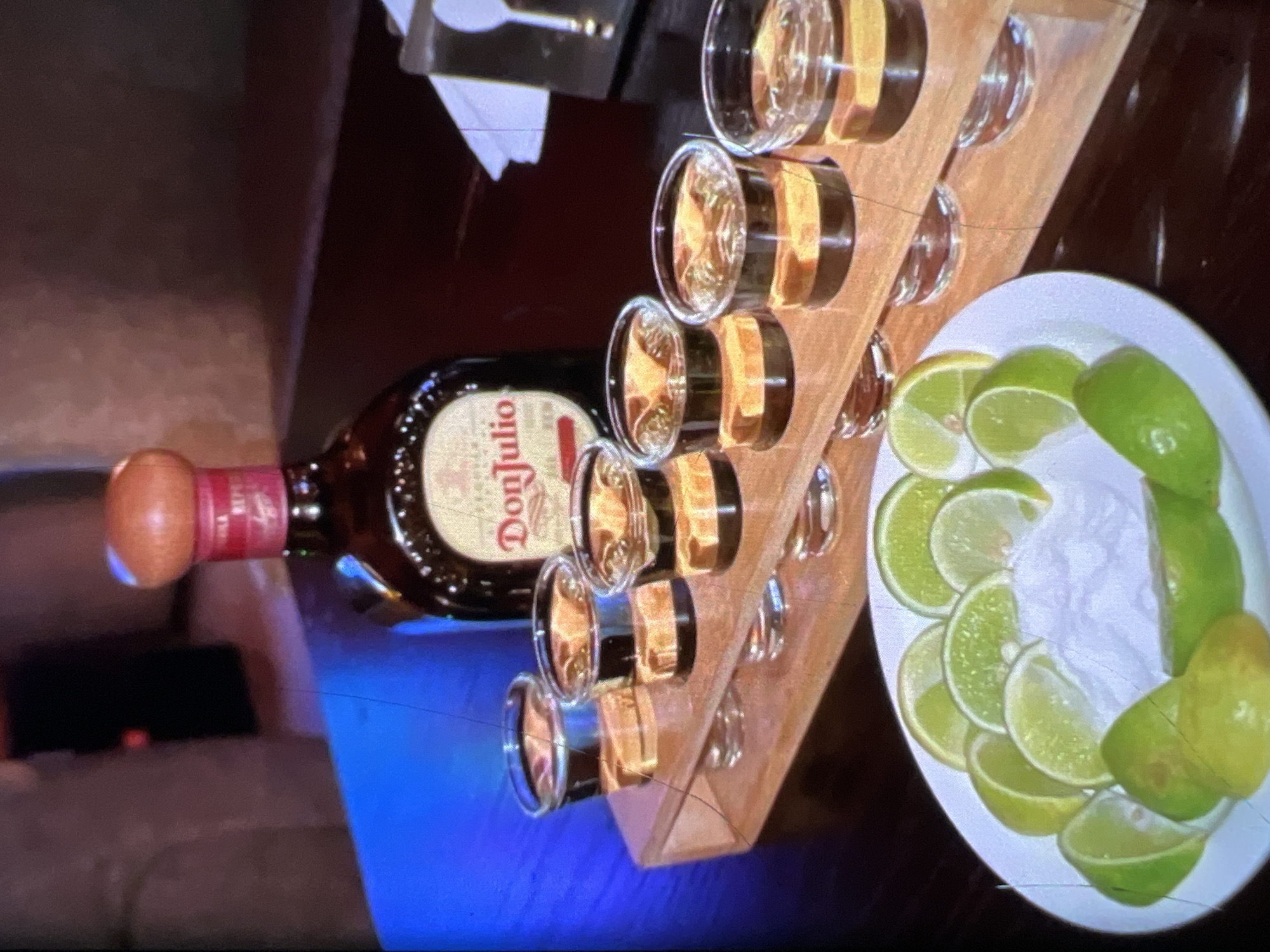 The image shows a serving of Don Julio tequila in shot glasses, arranged on a wooden holder. A bottle of Don Julio tequila stands behind the shot glasses. In the foreground, a white plate is filled with lime wedges and salt. The scene appears to be set in a dimly lit indoor environment, possibly a bar or restaurant. The blue lighting in the background suggests a modern, atmospheric setting, which is consistent with some high-end establishments in Lagos, Nigeria. There are no people visible in the image.