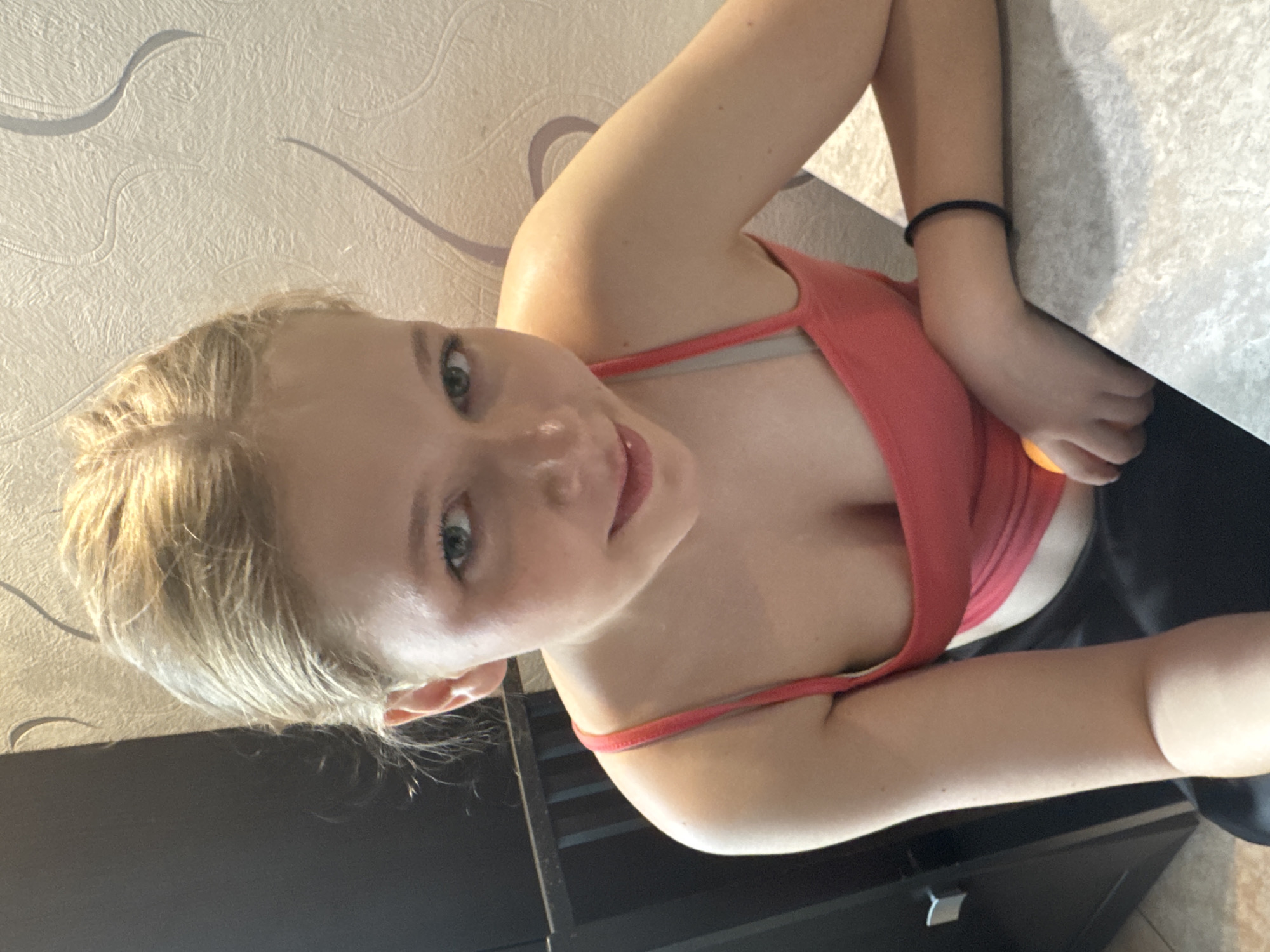 A young female individual is depicted in an indoor residential setting, appearing to be a teenager or young adult. She has blonde hair and light-colored eyes, looking directly at the camera with a slight smile. She is wearing a coral-pink tank top, under which a grey strap is visible on her left side, and dark-colored bottoms. A black bracelet or hair tie is present on her right wrist. Her left hand, partially visible, holds a small, spherical orange object, resting on a light-colored, speckled countertop or table. To her left, a dark brown or black cabinet with a silver handle is visible. To her right, the wall is covered in a light-colored, textured wallpaper featuring subtle, abstract wavy patterns. The scene takes place in a residential interior, located in the city of Perm, Russia.