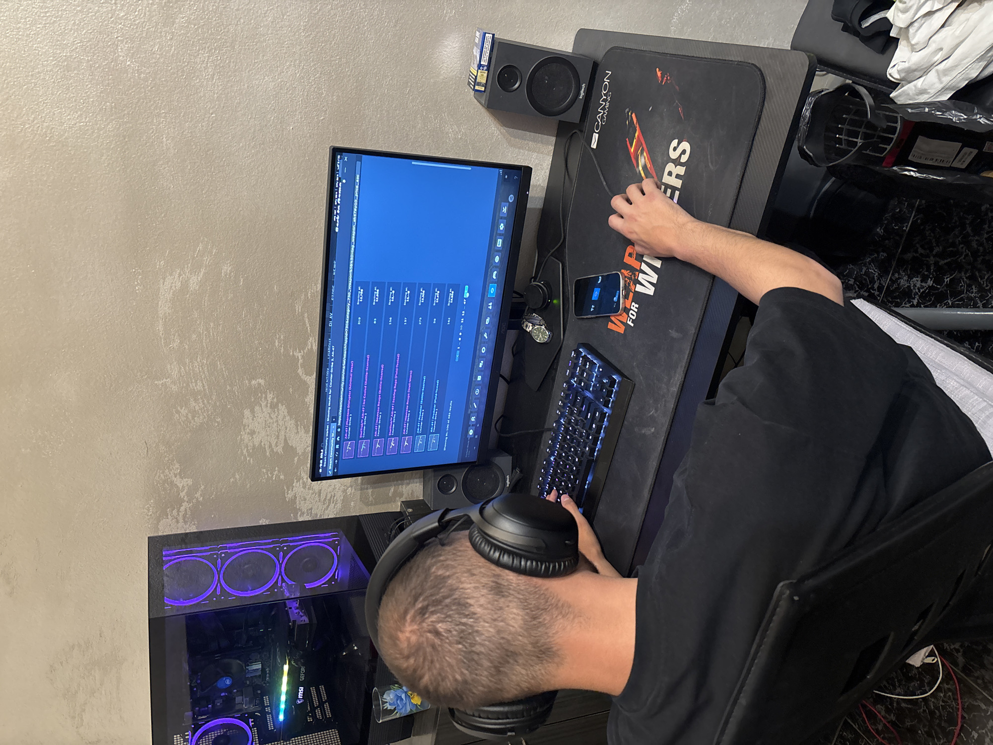 A man is sitting at a computer desk, wearing black headphones, and typing on a keyboard. A computer monitor displays what appears to be a search page, and a black speaker is to the monitor's left. A black mousepad with the words "FOR WINNERS" is to the monitor's right. A black computer tower with illuminated fans is on the floor to the left. A smartphone is resting on the desk next to the keyboard. The location in the city of Plovdiv, Bulgaria, is not determinable from the image.