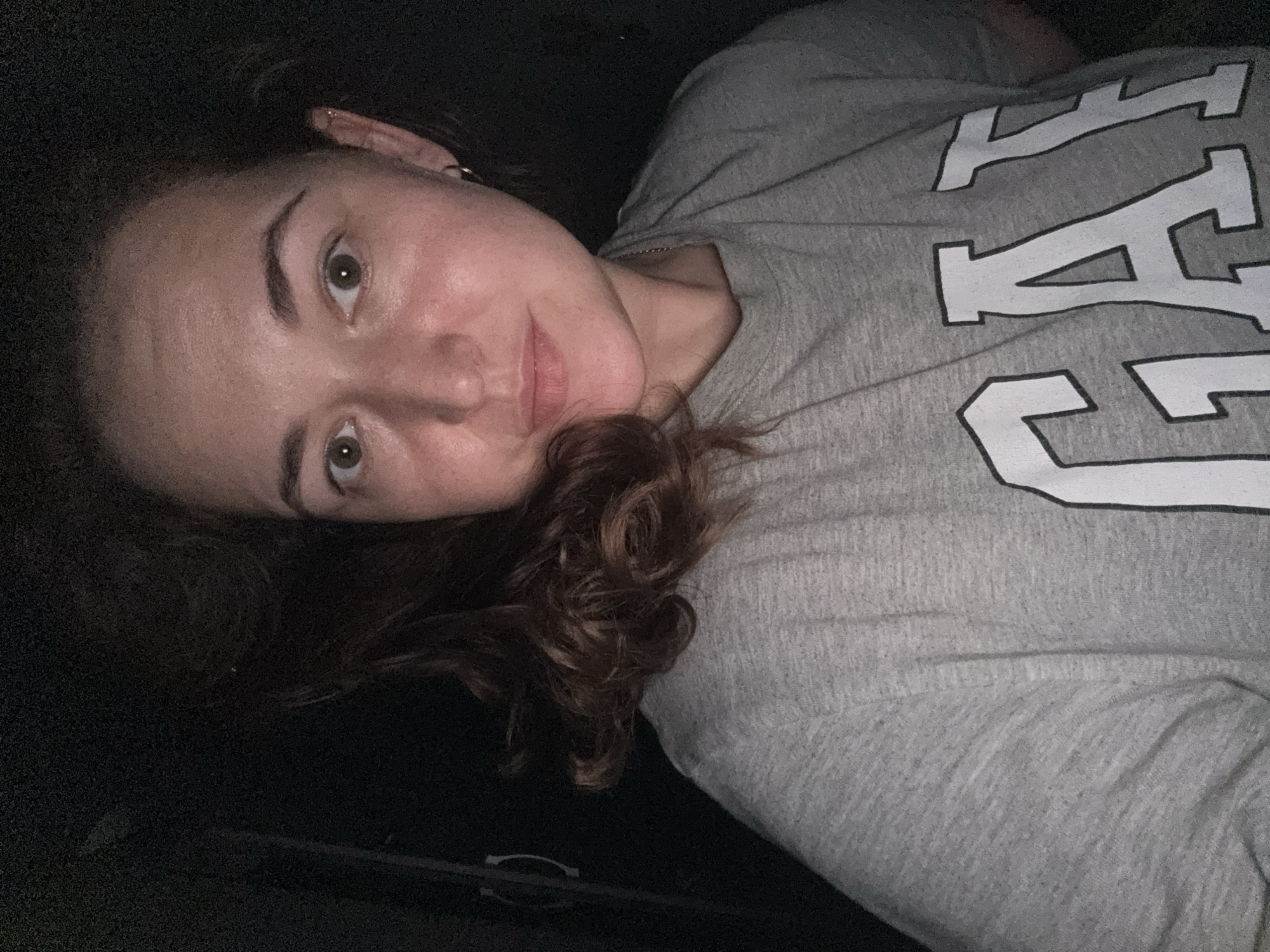 A medium shot captures a woman from the chest up, looking directly at the camera. She has light skin and is wearing a grey t-shirt with the word "GAP" printed in large white letters. Her medium-length, dark brown hair is styled loosely, with some strands falling around her face. The background is dark and indistinct. The image appears to be taken indoors with artificial lighting.