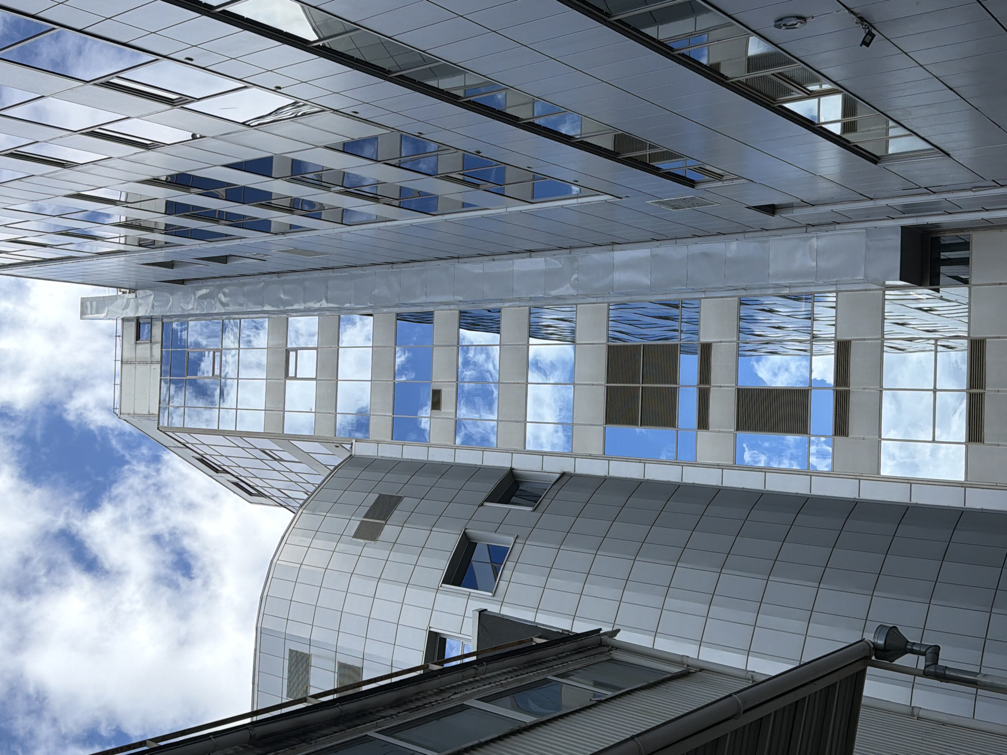 The image depicts a low-angle view of modern architecture, showcasing a cluster of buildings in Saint Petersburg, Russia. Dominating the frame are several structures featuring reflective glass facades, mirroring the sky with scattered clouds. The arrangement of the buildings creates a dynamic perspective, with different heights and shapes contributing to a contemporary cityscape.
Specifically, the buildings exhibit a mix of straight lines and curved surfaces. Some are adorned with a grid-like pattern of windows, while others have a smooth, curved exterior. The color palette is predominantly silver and gray, with the glass surfaces reflecting various shades of blue from the sky.
In the foreground, there is a partial view of another building with a visible rooftop and drainage system. The overall impression is one of a modern, urban environment, captured from an upward-facing perspective.