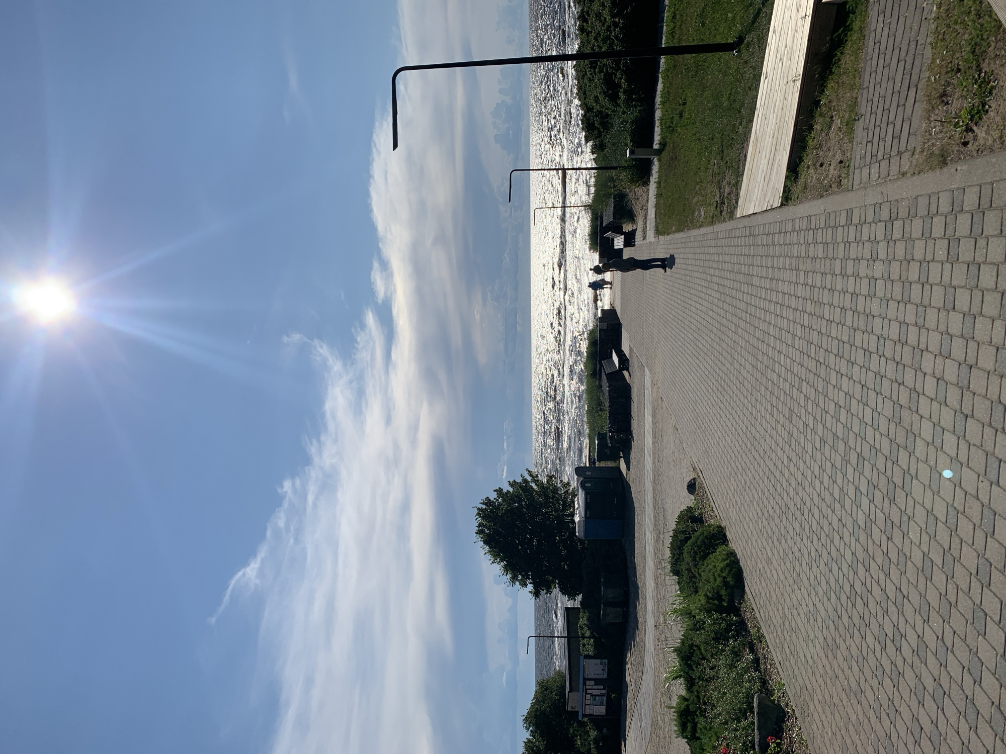 A bright sun shines in the upper right of a clear blue sky with scattered white clouds. A paved walkway leads away from the viewer towards a sparkling sea, where waves crash against the shore. On the left side of the walkway are bushes, trees, and several structures, including what appear to be portable toilets and a kiosk. Several people are walking on the walkway, heading towards the sea. There are also benches along the walkway. The environment is an outdoor public space, likely a beach or promenade. The location is identified as Saulkrasti, Latvia.