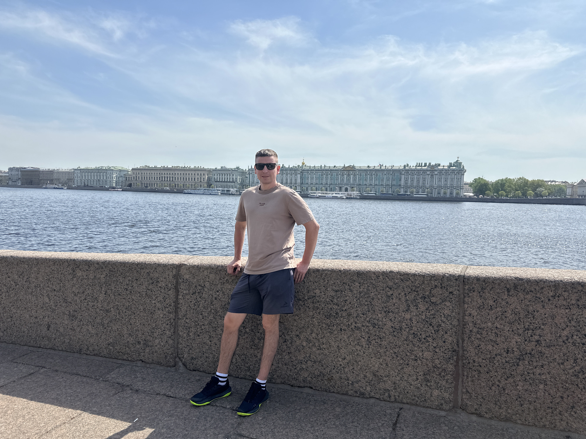 A medium shot presents a man in sunglasses, a beige t-shirt, and blue-gray shorts, leaning against a gray stone wall. He's also wearing black athletic shoes with neon green soles and white socks. The background reveals a wide river and several large, ornate buildings, typical of Saint Petersburg, Russia. Based on the architectural style and location along the Neva River, the buildings appear to include the Winter Palace (State Hermitage Museum). The sky is light blue with scattered clouds.