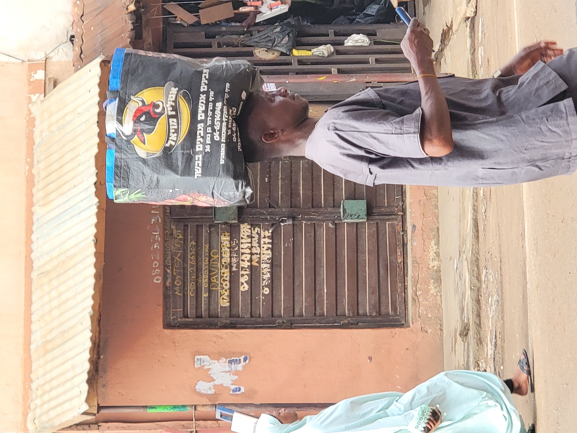 A man balances a large bag with Hebrew lettering and a stylized logo of a cow's head on his head in an outdoor setting. The bag seems to be for a butcher shop. He is wearing a gray garment and holding a blue object. Behind him is a closed metal door with more information painted on it, including phone numbers. Part of another person in a blue garment is visible at the bottom of the frame. The environment appears to be a street in Maiburiji, Nigeria.