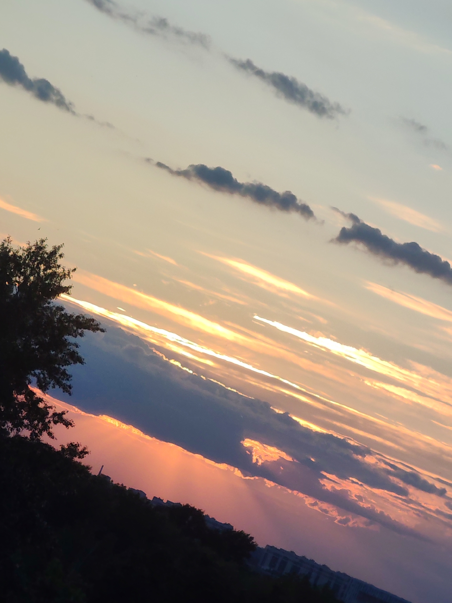 The image displays a vibrant and dramatic sunset or sunrise over Podolsk, Russia, captured with a diagonal tilt. The sky dominates the frame, transitioning from a pale blue-grey at the top to intense oranges, pinks, and purples closer to the horizon.
Long, wispy, dark grey clouds are scattered across the upper sky. Below these, a more substantial band of dark, dense clouds stretches diagonally across the mid-section, acting as a canvas for the setting or rising sun's light. Brilliant streaks of golden and orange light emanate from behind or below these clouds, illuminating their edges and creating glowing horizontal patterns across the sky. Patches of deep pink and purple hues are visible where the light filters through the atmospheric layers.
In the foreground on the left, a large, dark silhouette of a tree with visible branches and foliage stands out against the bright sky. Along the entire horizon, a dark silhouette of an urban or suburban landscape is visible, featuring numerous buildings and a continuous line of dense trees and foliage. A distinct, slender structure, possibly a tower or chimney, can be seen rising above the building silhouettes in the lower right portion of the frame. The scene powerfully captures the natural beauty and dynamic light of dusk or dawn. No visible text is present.