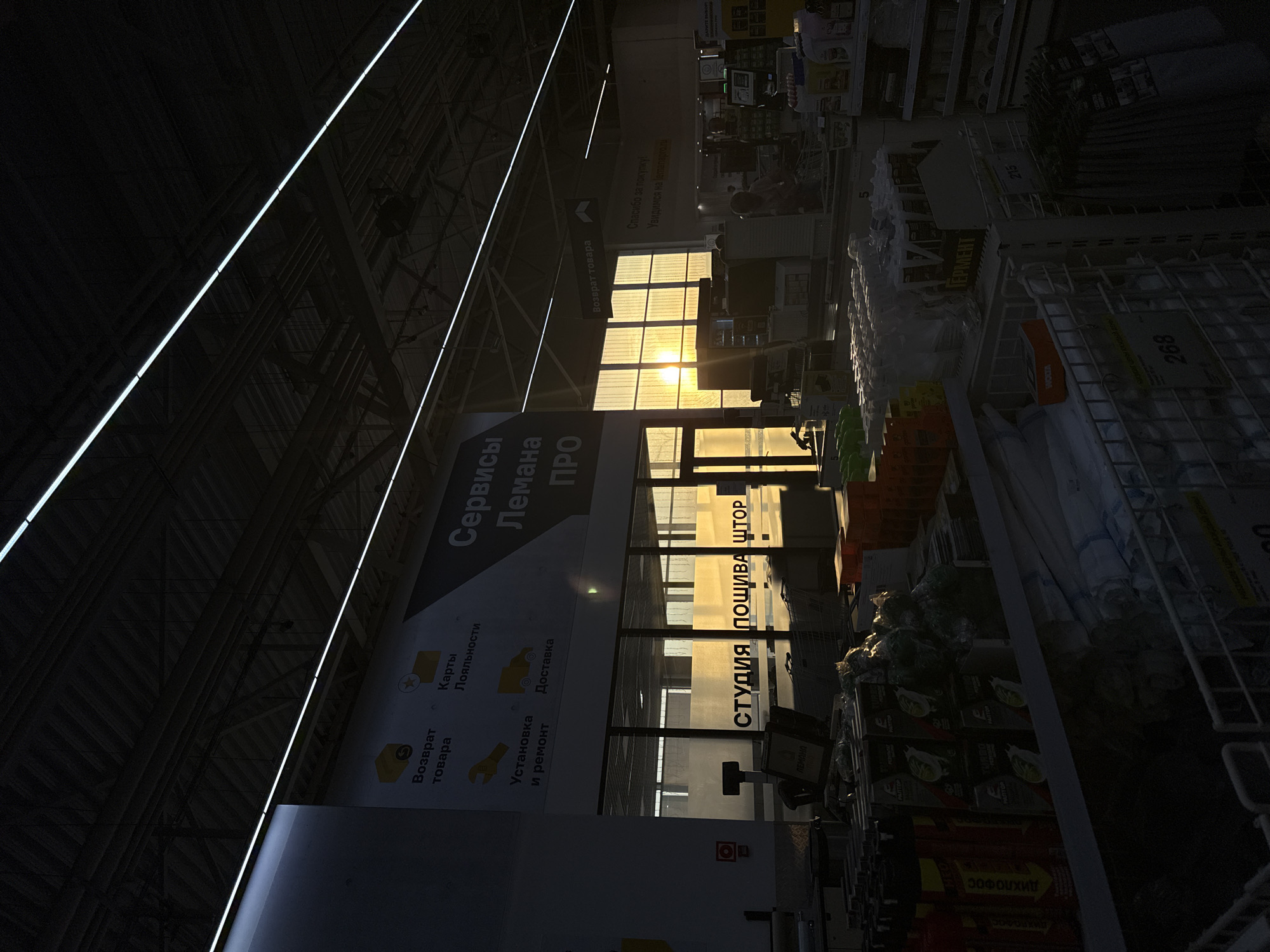 This is a photograph taken inside a large retail store, possibly a hardware or home improvement store, in the city of Yudinо, Russia. The setting appears to be a wide aisle with shelves stocked with various products, including what looks like cleaning supplies, aerosols, and rolled goods such as curtains or blinds. In the foreground, several shopping carts are visible, some empty and some partially filled.
The store's interior is dimly lit, with the primary light source coming from a large window or glass facade at the rear of the store, through which the setting sun is prominently visible. The sunlight casts a warm, golden glow across the scene, silhouetting some of the interior structures and creating a dramatic contrast with the darker areas. Fluorescent strip lighting runs along the ceiling.
Prominent signage in Russian is visible on the walls. One large sign reads "Студия пошива штор," which translates to "Curtain Sewing Studio." Another sign indicates "Сервисы Лемана ПРО," suggesting "Lemanа PRO Services." Other signs mention "Возврат товара" (Return of goods), "Карты Лояльности" (Loyalty cards), "Установка и ремонт" (Installation and repair), and "Доставка" (Delivery).
The scene captures a transitional moment of the day, with the last rays of sunlight illuminating the interior of the store. There are no clearly identifiable individuals in focus, although figures are faintly visible in the background near the checkout area, suggesting the store may still be open. The overall atmosphere is one of a typical commercial environment during a striking sunset.