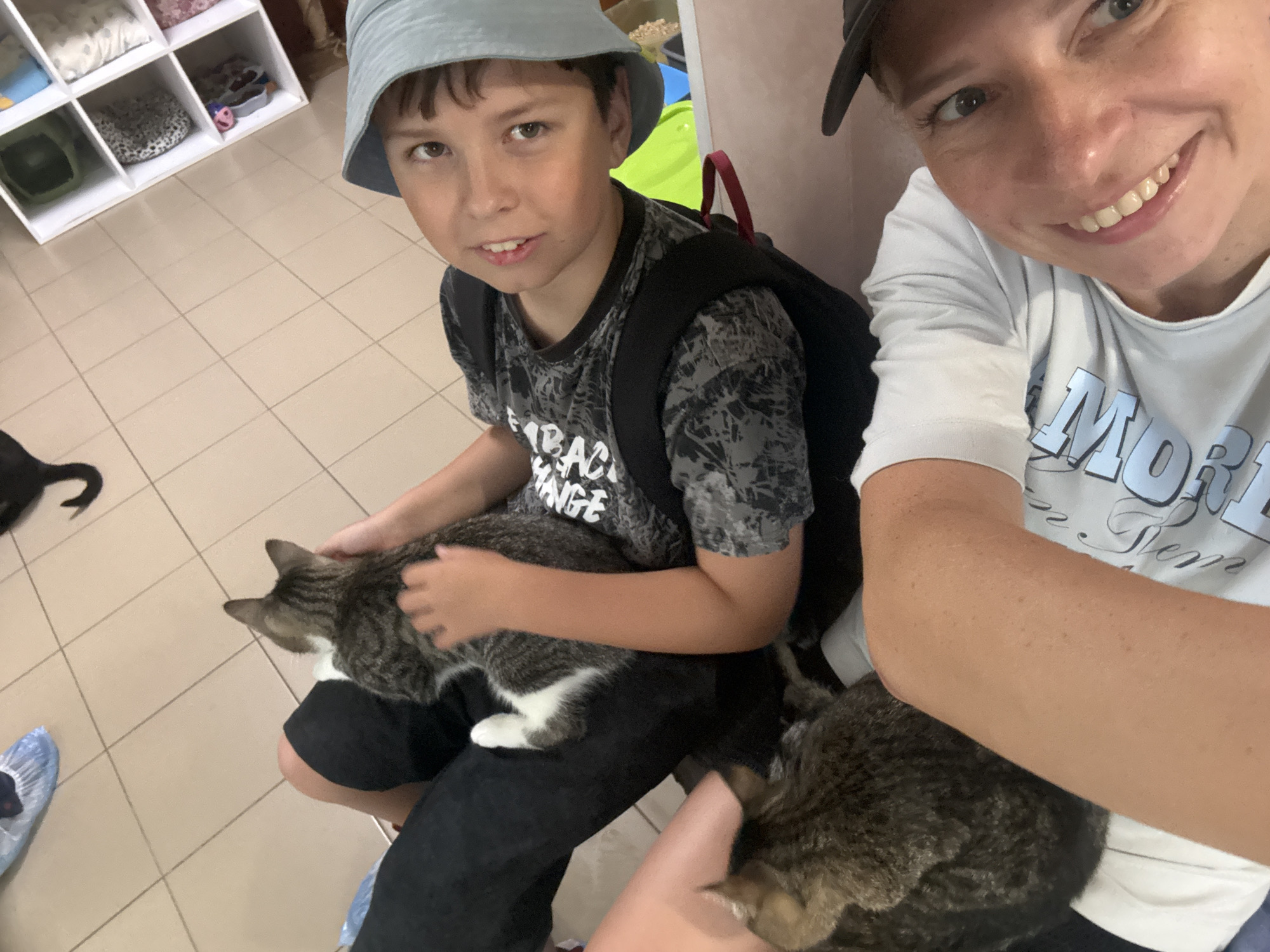 The image captures a close-up selfie-style shot featuring two individuals and three cats. On the left, a young boy wearing a light blue bucket hat and a dark grey t-shirt with a patterned design is seated. He is looking towards the camera with a neutral expression. He has a black backpack on. In his lap, he is holding a tabby and white cat. To the right, an adult woman, possibly the boy's mother, is also looking at the camera with a wide smile. She is wearing a light grey t-shirt with blue text that is partially visible. Another cat, primarily grey and black tabby, is resting on her lap and arm. A third cat, a black feline, is partially visible in the background on the left. The setting appears to be indoors, with tiled flooring and a white shelving unit in the background stocked with various items, some of which appear to be cat beds or supplies. The overall atmosphere is casual and affectionate, with the people interacting with the cats. There is no information available to determine the specific location within the city of Petrozavodsk, Russia.