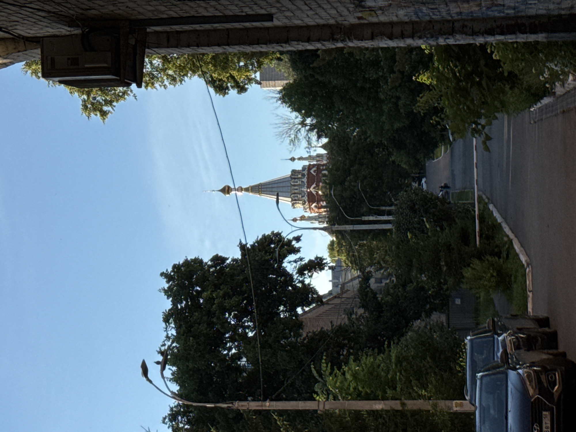 The image captures an outdoor scene, likely in Izhevsk, Russia, under a clear blue sky, suggesting it is daytime with fair weather. The perspective is from a low vantage point, looking upwards and outwards, framed by an overhead brick structure and a dark, horizontal element that could be a cable or overhang.
The primary subjects are a collection of cars parked along a road on the right side of the frame. These appear to be modern vehicles, with at least one black SUV prominently visible, its license plate showing Cyrillic characters and numbers (partially obscured). The road itself is paved and curves slightly.
In the background, amidst lush green trees and foliage, the domed roof and spires of a building, likely a church or cathedral, are visible. The architecture suggests a Russian Orthodox style, with multiple ornate domes and intricate decorative elements. Further back, other buildings are partially visible, peeking through the trees, indicating a developed urban or semi-urban setting.
On the left side of the frame, a large tree with dense foliage dominates the mid-ground. A streetlamp with two lamps, one of which appears to be a bud-like shape, is also visible in the lower left, contributing to the urban streetscape. A utility box or electrical equipment is mounted on a brick wall in the upper left corner.
The scene appears peaceful and ordinary, with no specific event taking place. The lighting suggests it is a sunny day. There are no people clearly discernible in the image, though the presence of parked cars implies human activity in the vicinity.