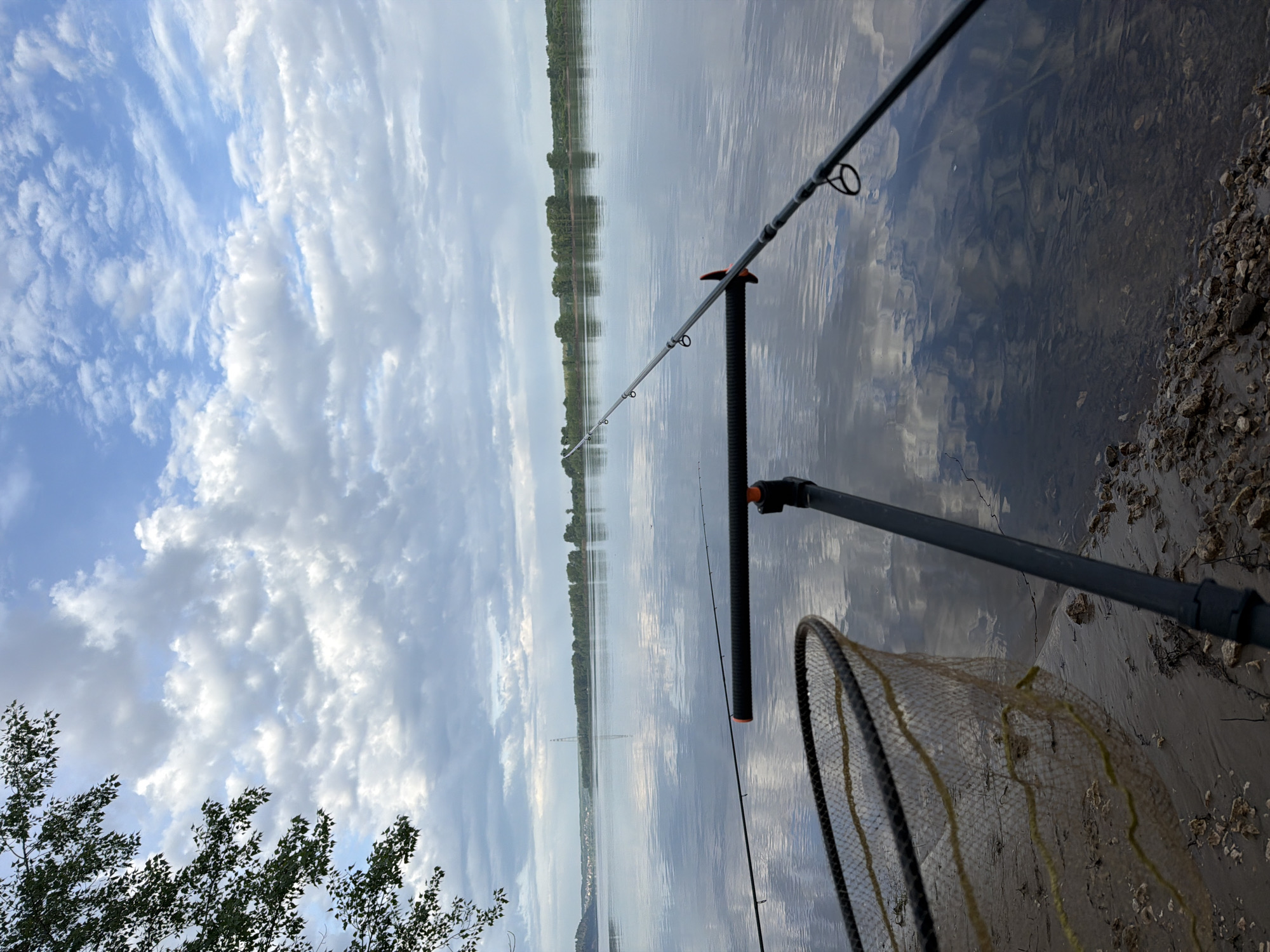 The image captures a fishing scene on the Kama River in Chaikovsky, Russia. A fishing rod is positioned on a rod rest, with the river reflecting the sky. A fishing net lies on the bank. The sky is partly cloudy. In the distance, the opposite shore of the river is visible.