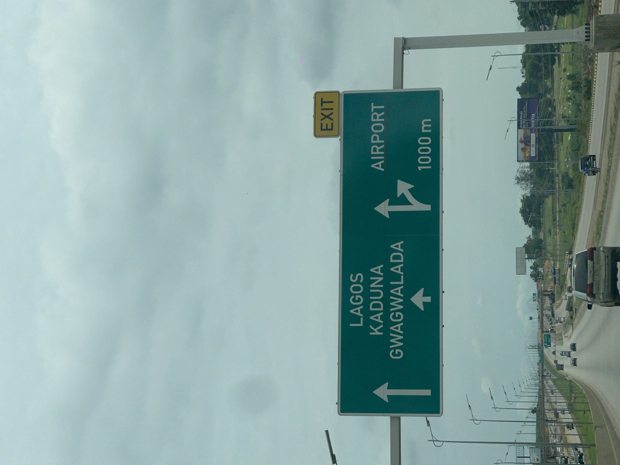 An eye-level, medium shot captures a green highway sign with white lettering and arrows, indicating destinations and distances. The sign reads "LAGOS KADUNA GWAGWALADA" with an upward arrow on the left, and "AIRPORT" with a right-turn arrow and "1000 m" on the right. Above the green sign, a yellow rectangular sign reads "EXIT." The sign is positioned over a multi-lane highway with vehicles in motion. The sky is overcast with grey clouds. To the right of the green sign, a billboard is visible. The highway is bordered by grassy verges and trees. The scene is located on a highway leading towards the Immigration Quarters in Nigeria.