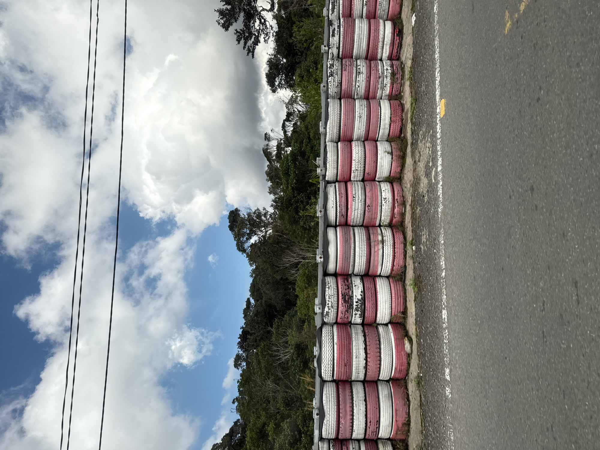 The image depicts a roadside scene featuring a barrier constructed from stacked tires. The tires are predominantly red and white, arranged in multiple rows and forming a substantial wall. Behind the tire barrier, a dense forest with a variety of trees stretches across the background. The sky above is partly cloudy, with patches of blue visible between large, white cumulus clouds. Power lines are strung across the upper portion of the frame, running horizontally above the trees. The foreground shows an asphalt road with a white line marking. The overall impression is one of an outdoor location, possibly a race track or a road along a wooded area, in Nam Khánh Vĩnh, Vietnam, given the provided context.