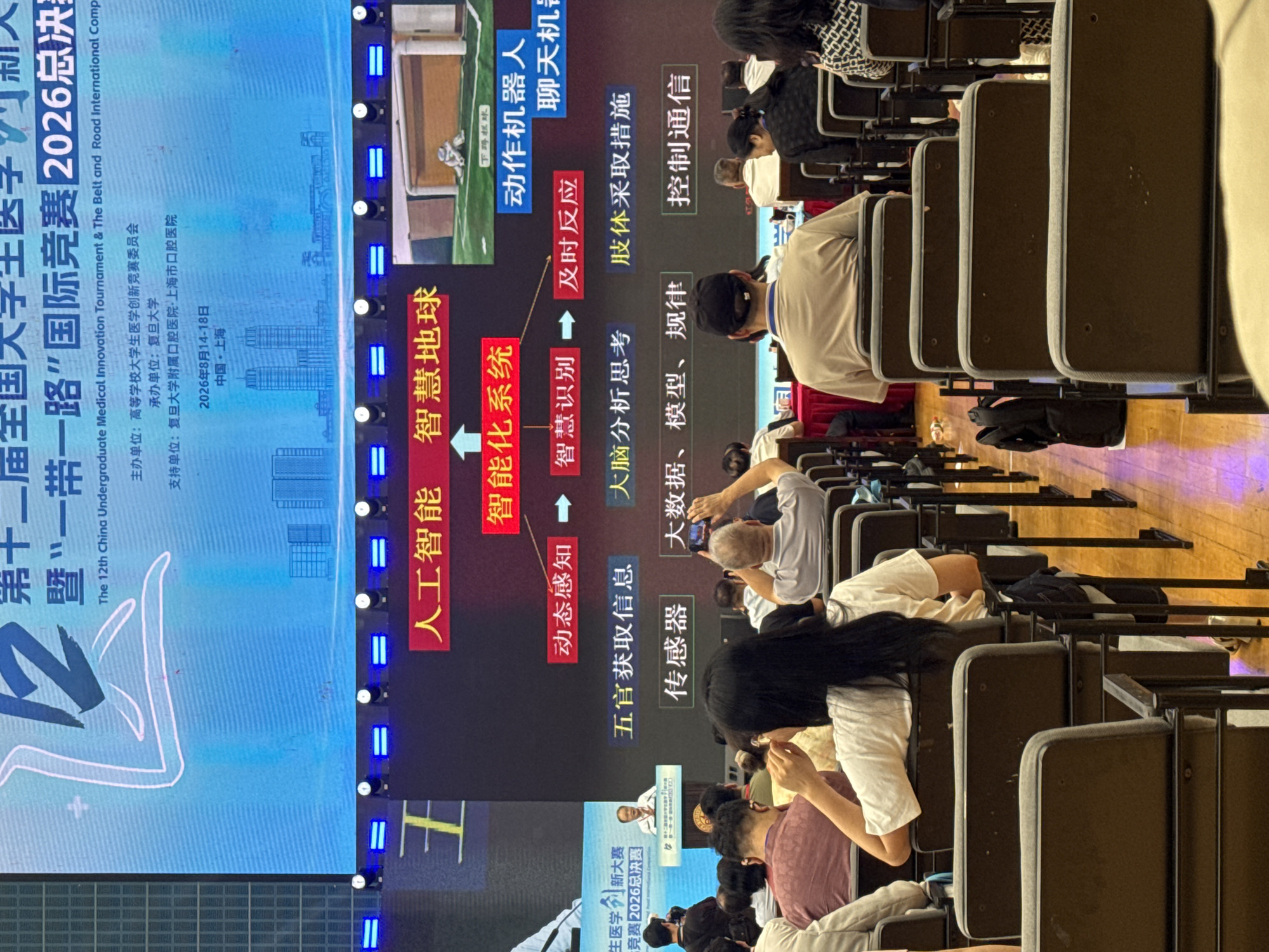 The image captures a presentation taking place in a lecture hall or auditorium. The foreground shows the backs of several audience members seated in dark brown chairs. The audience members are diverse in age and gender, with some facing forward and others turned slightly. Many are looking towards a large screen that dominates the background.
The screen displays a graphic flowchart with Chinese text. At the top, it shows "The 12th China Undergraduate Medical Innovation Tournament & The Belt and Road International Competition 2026 Finals." Below this, there are various terms related to artificial intelligence, such as "Artificial Intelligence," "Smart Earth," "Intelligent System," "Dynamic Perception," "Intelligent Recognition," and "Timely Response." Further down, there are phrases like "Sensory Input," "Big Data, Models, Rules," "Brain Analysis and Thinking," "Body Response," and "Control Communication." The screen also shows a video clip of a robot playing soccer.
A speaker is visible at a podium on the left side of the screen, addressing the audience. The room itself appears to be a modern lecture hall, with the chairs arranged in rows facing the screen. The lighting suggests an indoor event. The overall atmosphere is that of an academic or technological conference or competition. The mention of "Yangpu District, China" in the prompt suggests the location of this event.
