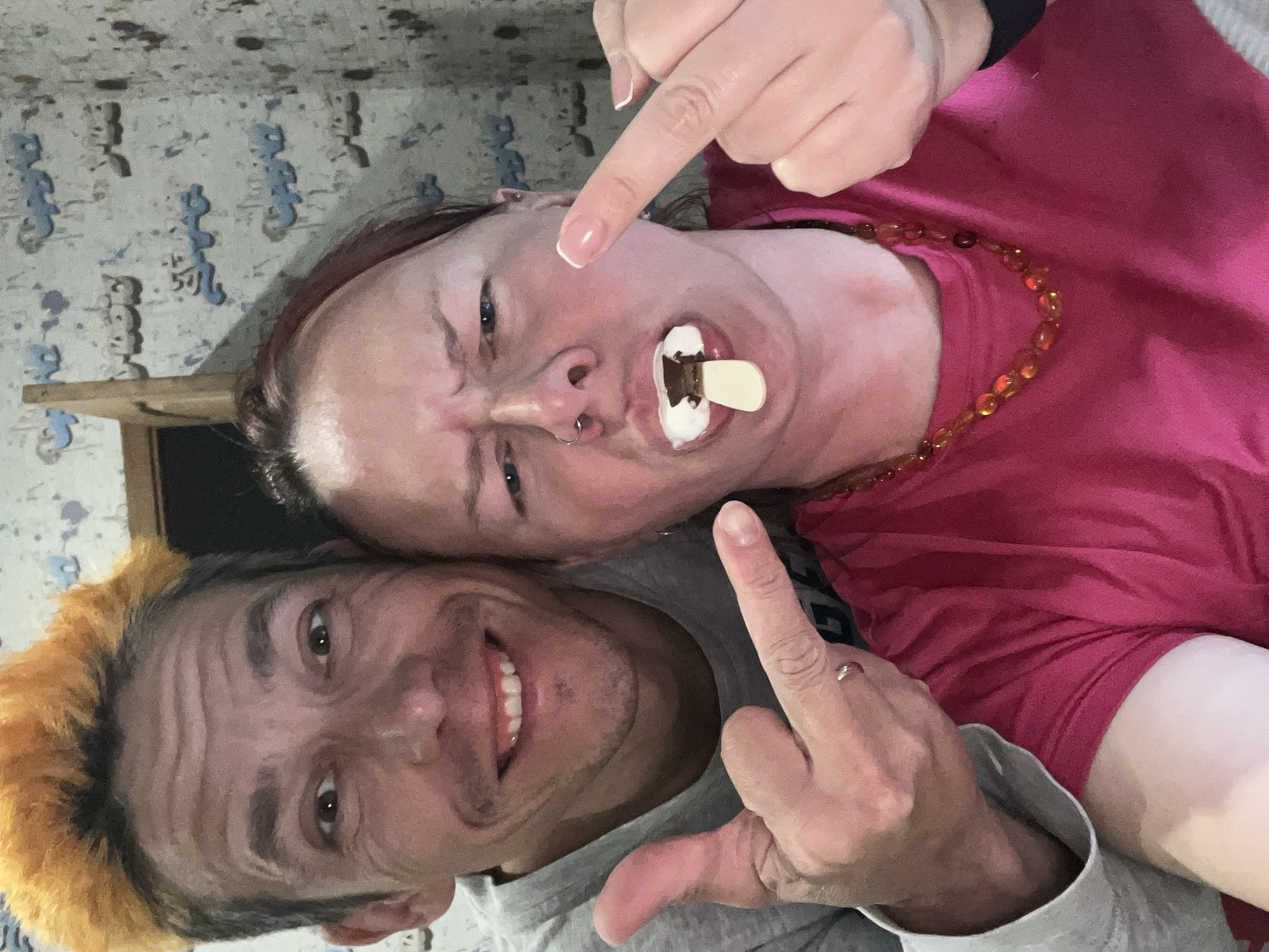 Two people are in the image. The person in the top right is wearing a pink shirt and amber-colored beaded necklace. They are sticking out their tongue with a popsicle stick in their mouth, with something white on the stick. They are grimacing and pointing their index finger down toward their mouth. The person in the bottom left is wearing a gray shirt with yellow-tipped hair. They are smiling. Both people are extending their middle finger. The background is a wall with a blue and white design. There is also a small wooden shelf visible. There is no information about the location in the city of Chaikovsky, Russia.
