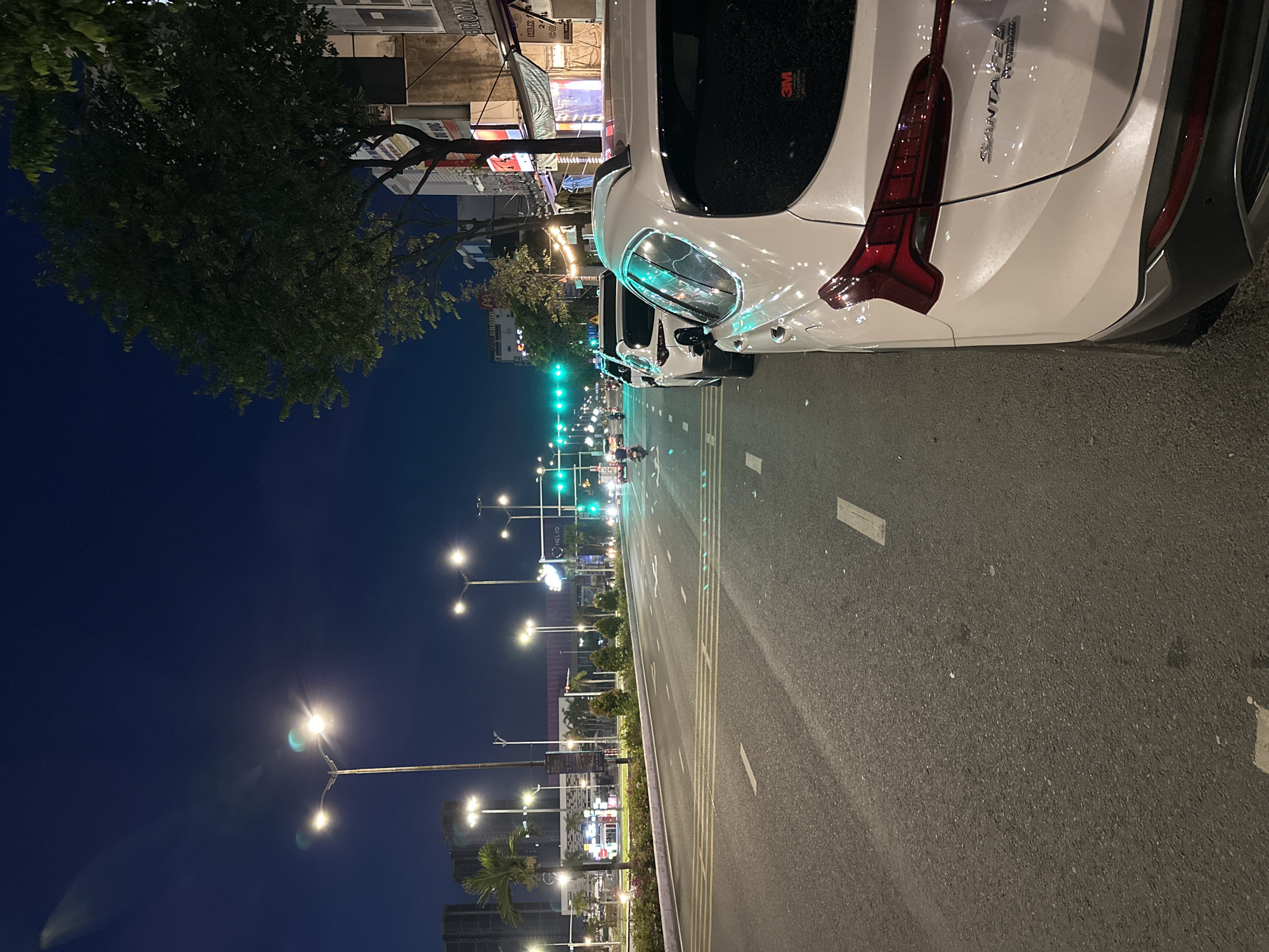 The image depicts a street scene at night in Da Nang, Vietnam. The viewpoint is from the side of the road, looking down the street. In the foreground, a white SUV is parked on the right side of the road, its rear visible. The street itself is paved and has white and yellow lane markings. Across the street, there are more parked cars and moving vehicles, including motorcycles and a scooter. Streetlights illuminate the road, casting a warm glow. The sky is a deep blue, indicating it is nighttime. On the left side of the street, there are palm trees and buildings. The buildings on the right side appear to be shops or businesses, with awnings and signage visible. The overall impression is of a busy urban environment at night.