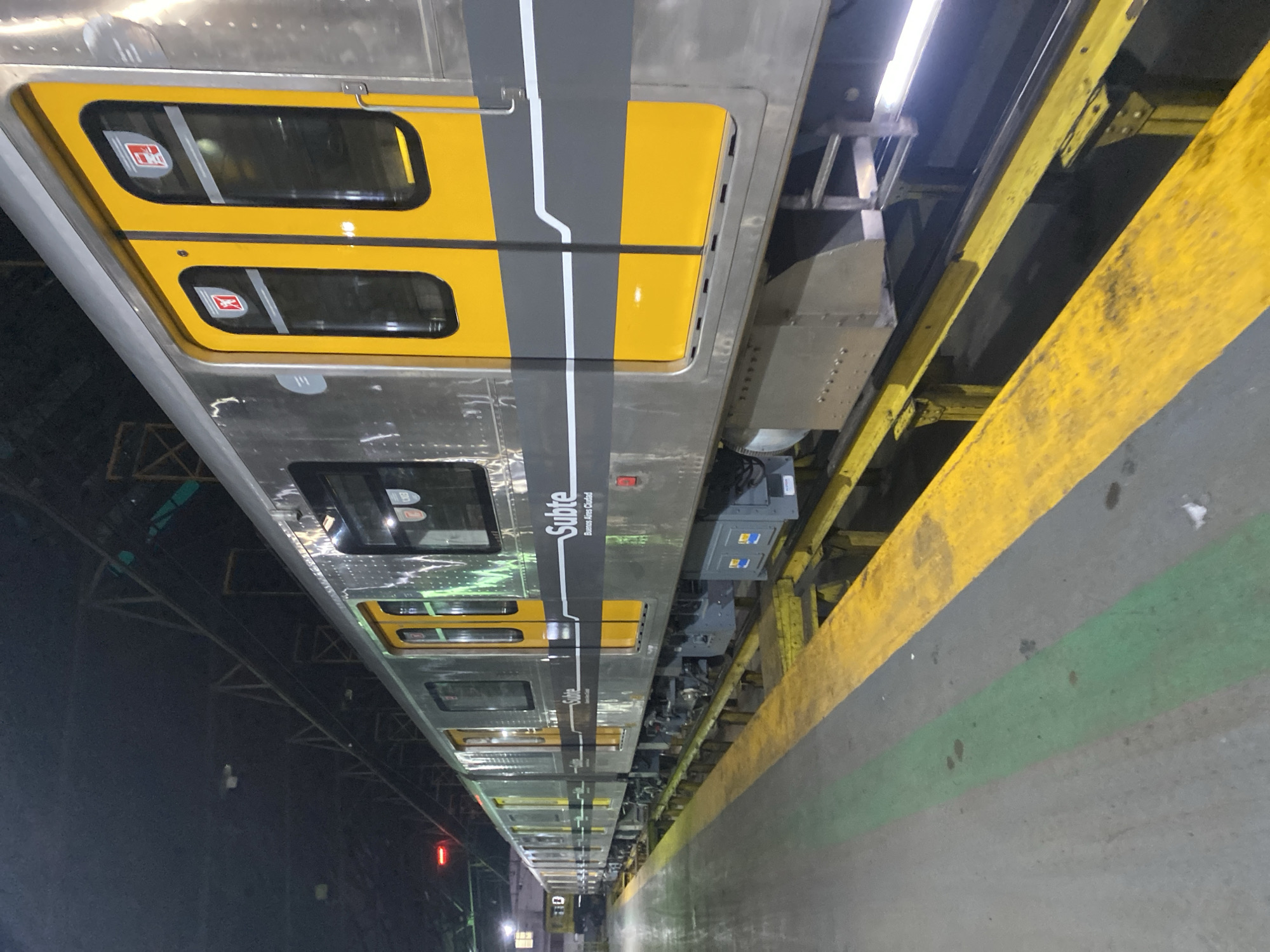 An overhead shot depicts a Subte (Buenos Aires Metro) train at a station platform, with the train's roof and side visible. The train is painted in a combination of silver and yellow, with a grey stripe running along its length that is labeled "Subte - Buenos Aires Ciudad." The yellow doors have rectangular windows. The platform edge is defined by a yellow safety line, and a green line is visible adjacent to it. The lighting is dim, suggesting an underground or depot environment. A small red signal light can be seen in the distance along the tracks. The perspective is from above and slightly to the side.