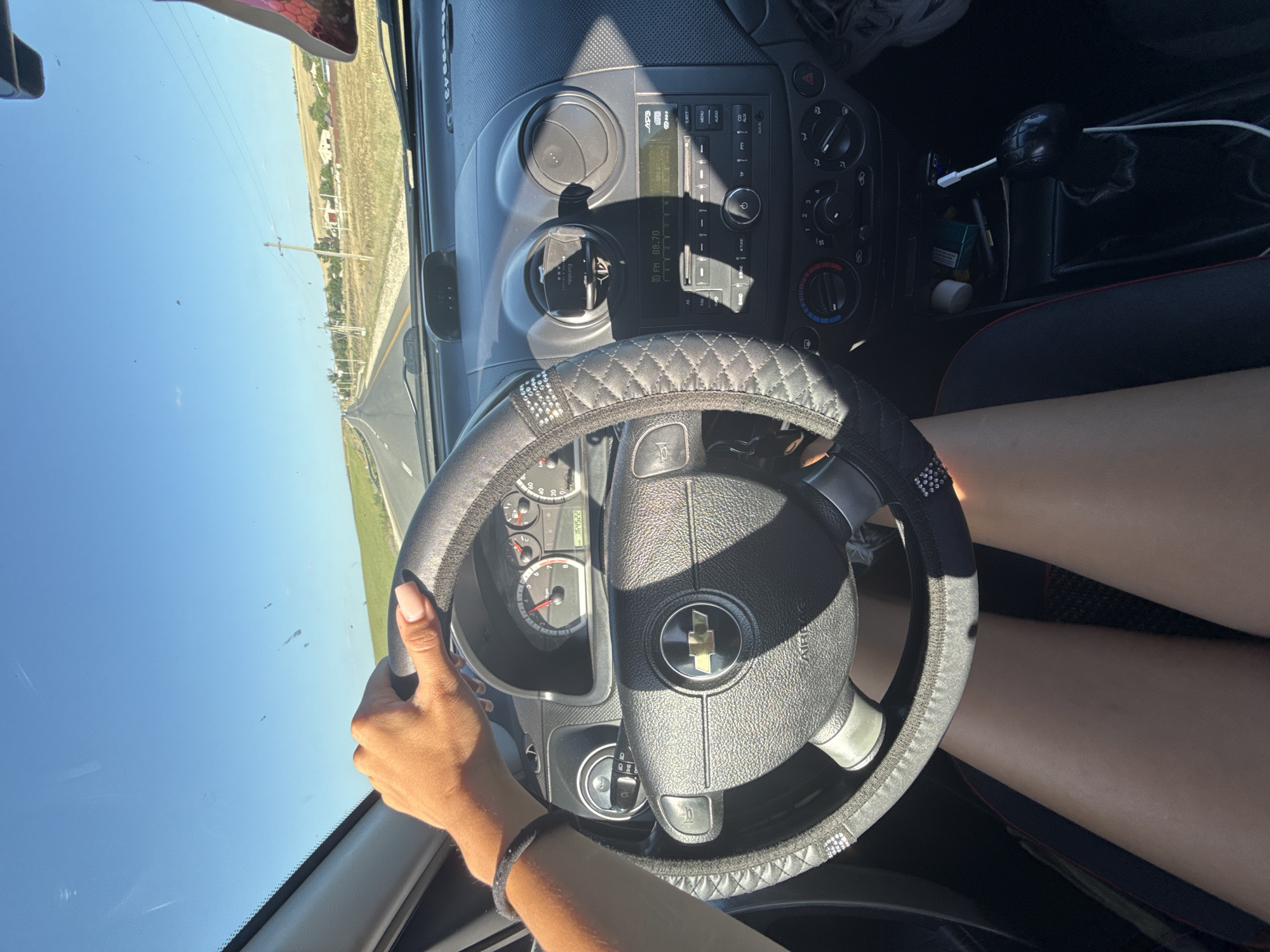 In the image, a person's hand is visible on the steering wheel of a car, which is being driven on a road. The interior of the car is seen, including the dashboard with a radio and air vents, and the gear shift. The driver's legs are also visible. The car is traveling through a rural area with fields on either side of the road. Power lines and some scattered houses can be seen in the distance. The sky is clear and blue, indicating a sunny day. The image appears to have been taken from the passenger seat or a similar vantage point inside the vehicle. The setting suggests a journey through the countryside of Comrat, Moldova, characterized by open fields, rural infrastructure, and a clear sky.