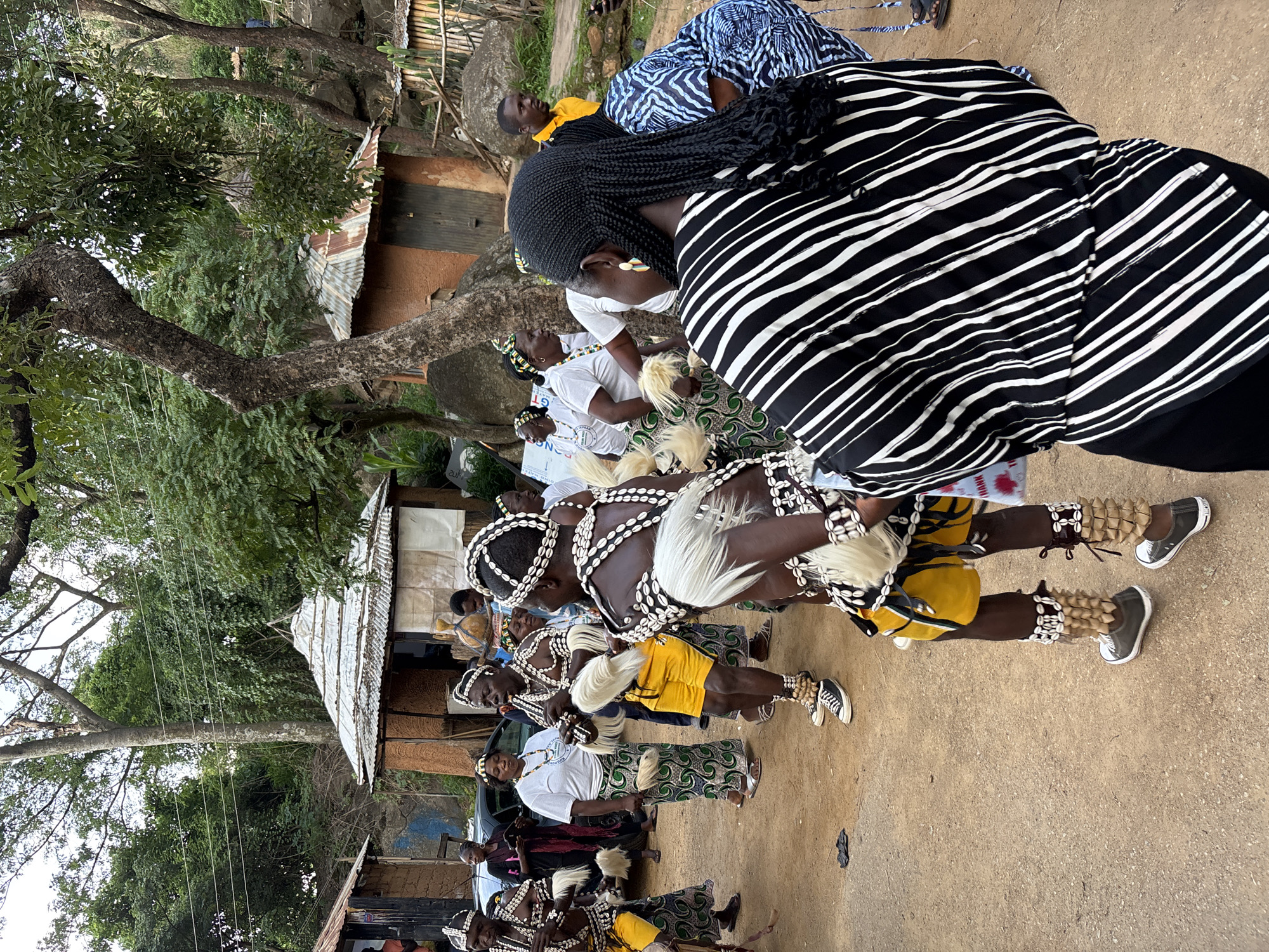 A group of people, some dressed in traditional attire with elaborate beadwork and white feather headdresses, are gathered in an outdoor, dusty area. The attire includes white beaded necklaces and armbands, yellow shorts for some individuals, and patterned skirts for others. Several individuals are wearing white t-shirts and skirts with a green and white geometric pattern. The environment appears to be a rural or semi-rural setting, possibly a village, with simple, earthy-toned structures in the background. Some of these structures have corrugated metal roofs. Tall trees with lush green foliage are visible, creating a canopy overhead. The ground is unpaved and sandy. In the foreground, a woman with braided black hair, wearing a black and white striped top, is the closest subject to the camera, with her back to the viewer. The scene suggests a cultural event or ceremony taking place in Jos, Nigeria.