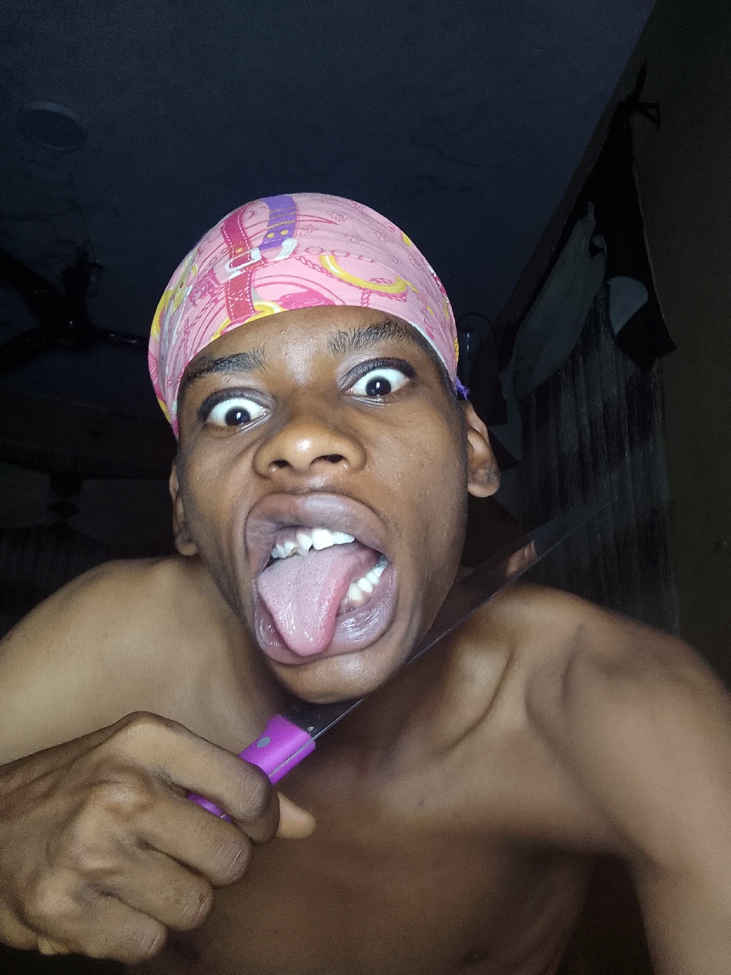 A man is pictured indoors. He is bare-chested and wearing a pink head covering. He is holding a knife with a purple handle to his neck. His mouth is open, and his tongue is sticking out. His eyes are wide. The background is dark. The location is Unknown, Unknown.
