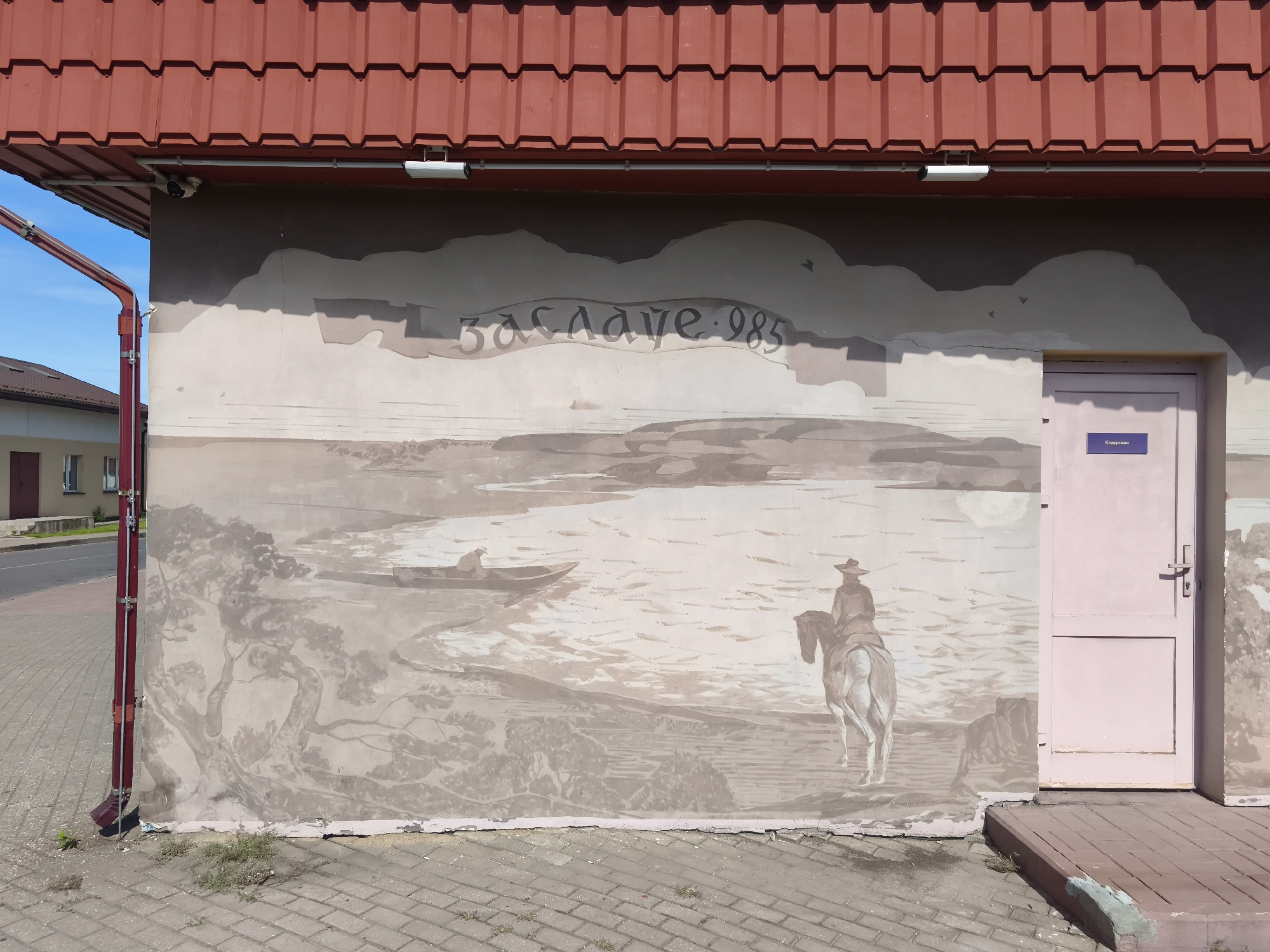 The image shows a mural painted on the side of a building in Zaslawye, Belarus. The mural depicts a landscape with a river, trees, a man in a boat, and a person on horseback. The mural is in sepia tones and has some cracks and peeling paint. Above the mural, the building has a red tiled roof and a downspout. A pink door is visible on the right side of the mural, with a sign on it that says "Касавая". The ground in front of the building is paved with bricks.