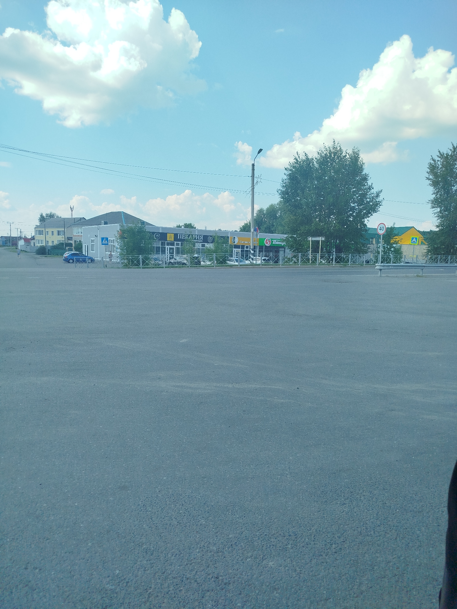 The image depicts a street scene in Zavodoukovsk, Russia, on a bright, cloudy day. In the foreground, a paved asphalt area, likely a parking lot or road, stretches towards a row of buildings. Behind the asphalt, a low metal fence separates it from a sidewalk and a grassy area. Beyond the fence, several buildings are visible, including a yellow two-story building on the left and a longer, lighter-colored building with a dark sloped roof in the center. The central building features a sign with Russian text that appears to read "ПЕКАРНЯ" (Bakery). Several cars are parked in front of this building. To the right of the bakery, a green building with a yellow triangular sign is visible, and further to the right, trees dominate the background. A lamppost stands to the left of the bakery. A speed limit sign indicating "40" is present on a pole to the right of the bakery. A few individuals are seen walking on the sidewalk in front of the buildings. The sky is blue with scattered white cumulus clouds.