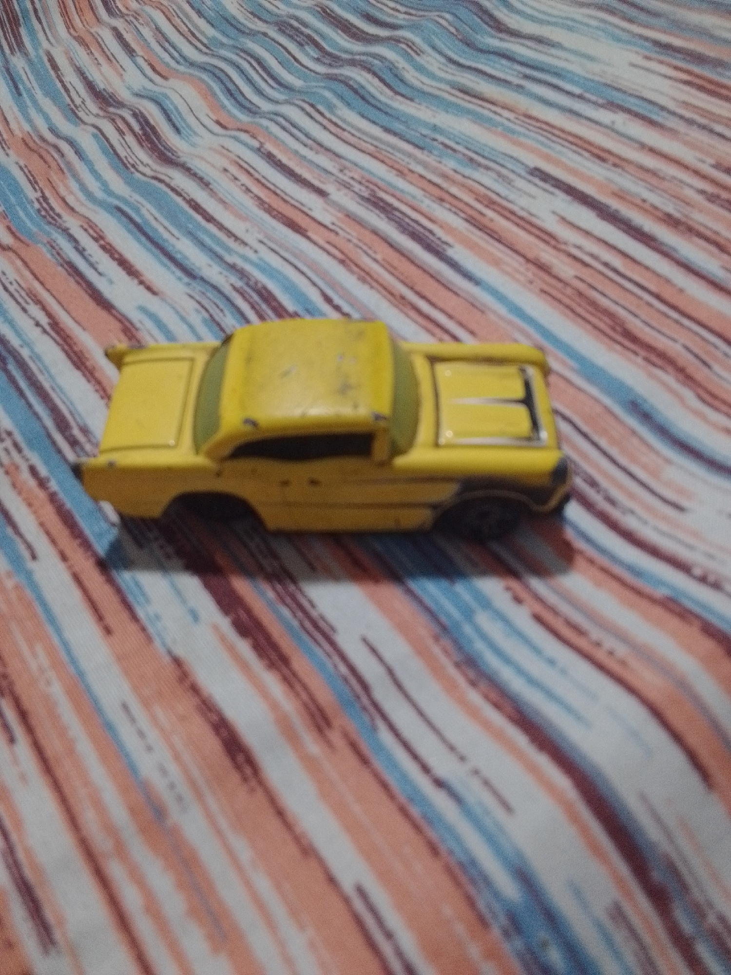 A yellow toy car is displayed on a striped fabric. The car appears to be a vintage model, possibly from the 1950s or 1960s, with a classic silhouette. It is lying on its side, facing towards the right of the frame. The fabric beneath the car has a pattern of diagonal stripes in shades of white, light blue, pink, and dark red. There is no information in the image to suggest a location in La Tagua, Venezuela.