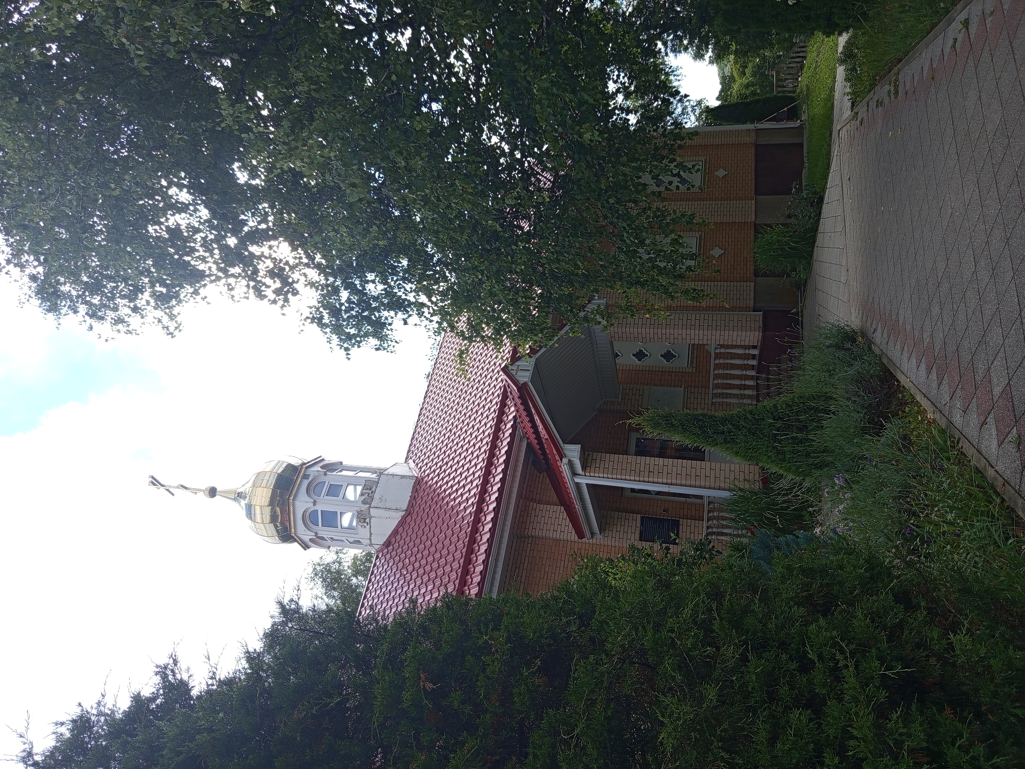 This image depicts an Orthodox church in Giaginskaya, Russia. The church is constructed of light-colored brick with a red tiled roof. It features a golden dome topped with a cross, and a white bell tower with arched windows. A paved walkway leads to the entrance, which is covered by a red awning. Evergreen trees and other greenery surround the church. The sky is partly cloudy.