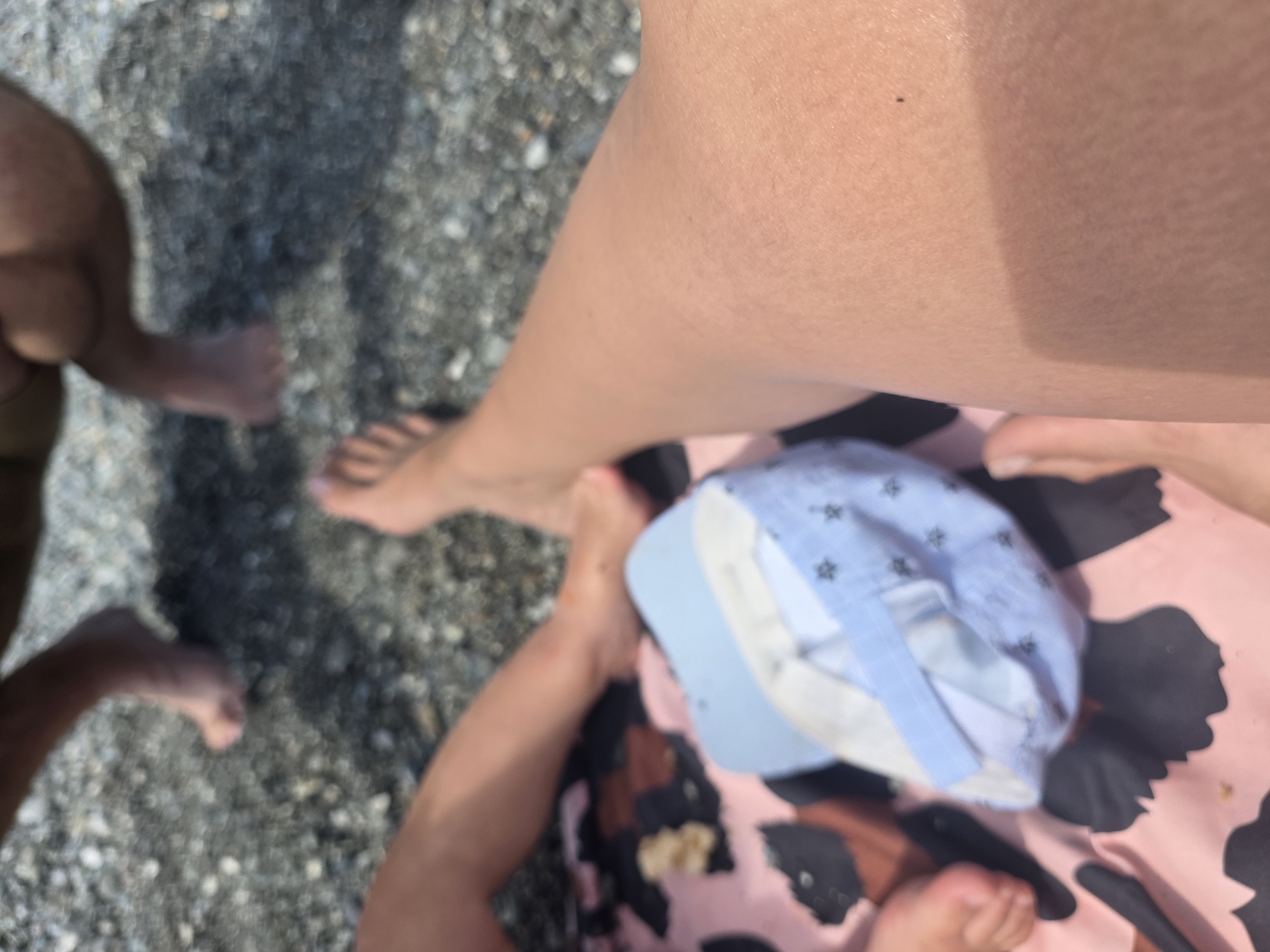 In Sochi, Russia, on a pebble beach, two individuals are seated. In the foreground, a person's legs are visible, one resting on a pink and black patterned blanket. A light blue baby hat with a star pattern is placed on the blanket. In the background, a second person's legs are also visible, with one foot resting on the pebbles. The environment is a typical pebble beach with a soft, diffused light suggesting daytime.