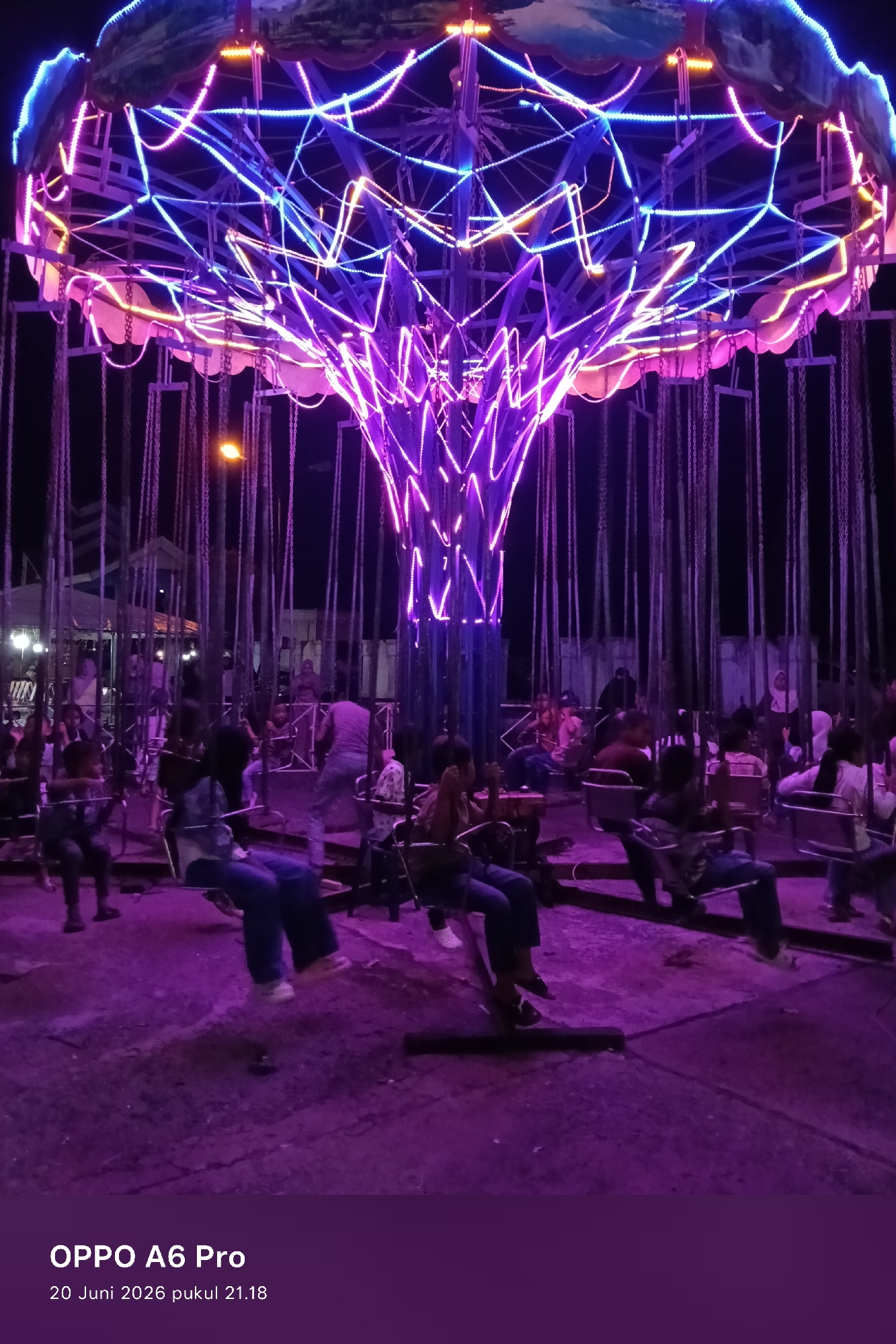 The image shows a swing ride at night, likely at an amusement park or fairground in Lhokseumawe, Indonesia. The ride is illuminated by bright purple and blue neon lights, creating a vibrant and festive atmosphere. Several people are seated on the swings, some appearing to be children and others adults. The ground beneath the ride is a rough, paved surface. The background is dark, suggesting the surrounding area is not well-lit or is further away. The photograph is overlaid with text indicating "OPPO A6 Pro" and the date "20 Juni 2026 pukul 21.18", suggesting it was taken with a mobile phone and includes a timestamp.