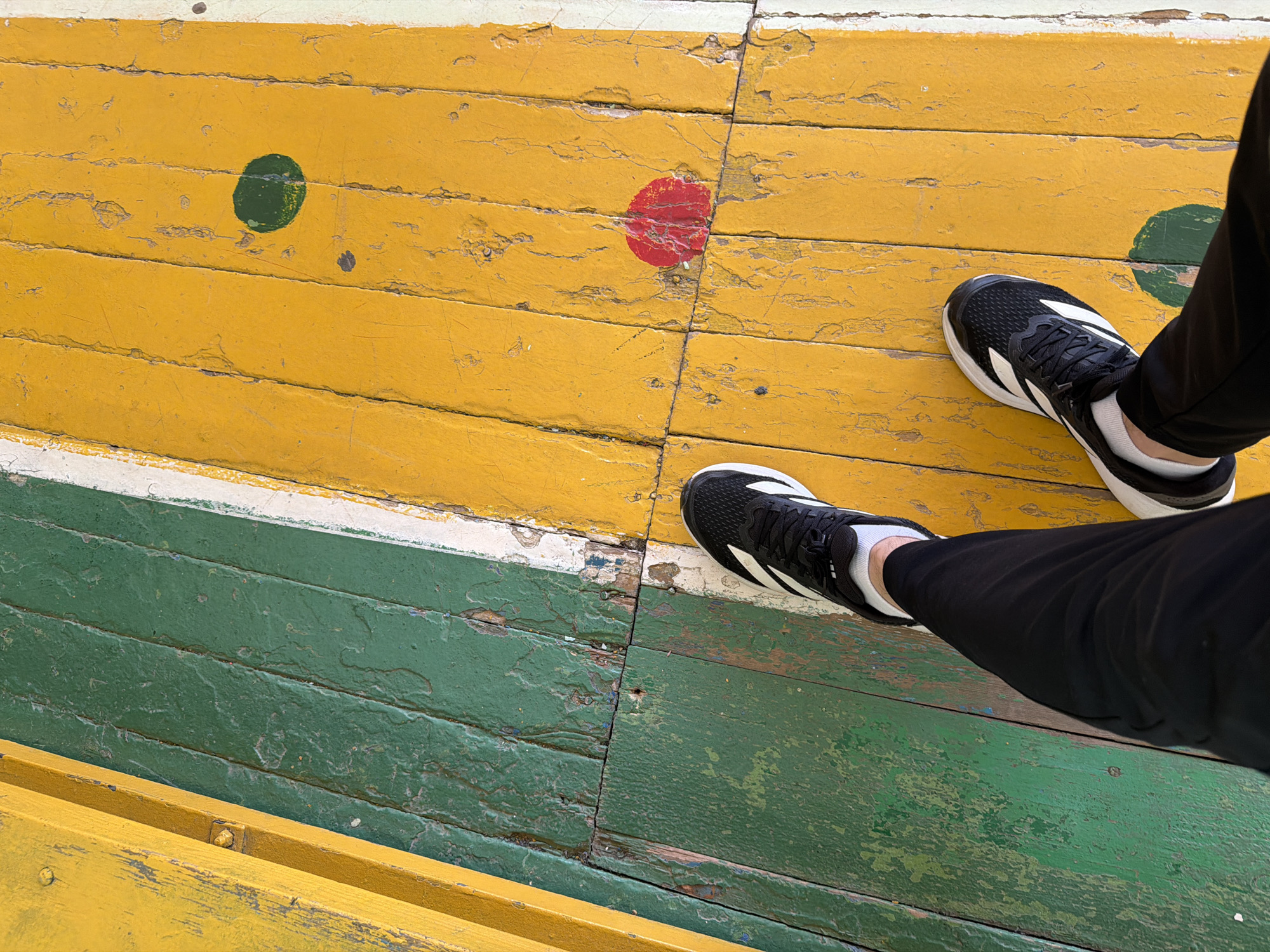 The image shows a person standing on a painted wooden surface. The surface is composed of planks arranged horizontally. The planks are painted in alternating stripes of yellow, green, and white. Several planks have circles painted on them, with one each in red and green. The paint is distressed, with signs of wear and chipping. The person is wearing black athletic shoes with white stripes and black pants. The location is not specified, and neither is the city of Izhevsk.
