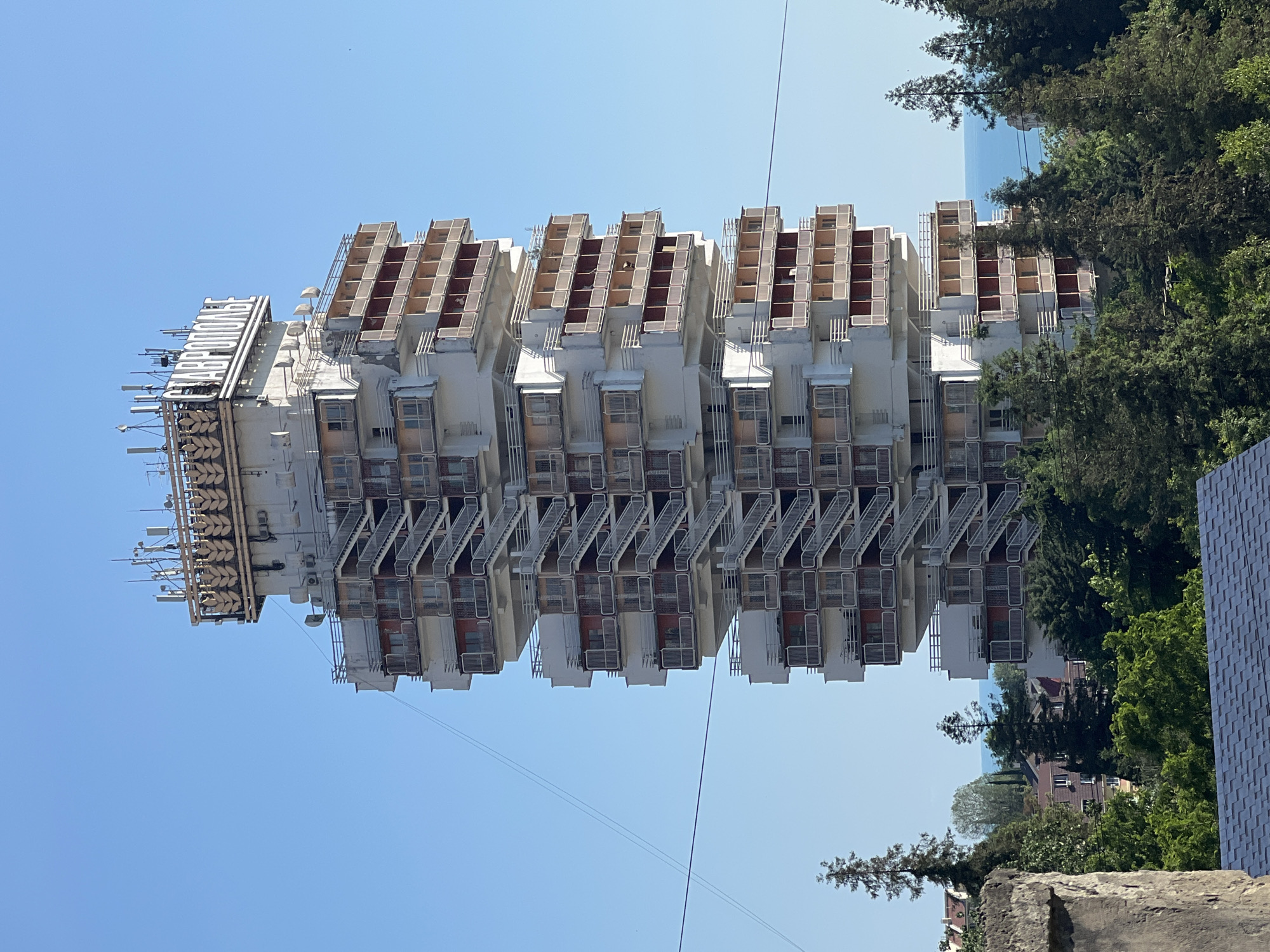 The image depicts a striking, Brutalist-style apartment building in Sochi, Russia, specifically the "Stavropolye" building. The building, characterized by its tiered balconies and external staircases, is situated amidst lush green trees, with a clear blue sky overhead and a hint of the Black Sea in the distance. The architecture of the "Stavropolye" building is its most prominent feature, showcasing a complex arrangement of cantilevered units and exposed stairwells, creating a unique and imposing silhouette. The top of the building features large wheat sheaf decorations and the building's name in Cyrillic lettering. The environment suggests a coastal city with abundant vegetation, typical of Sochi.