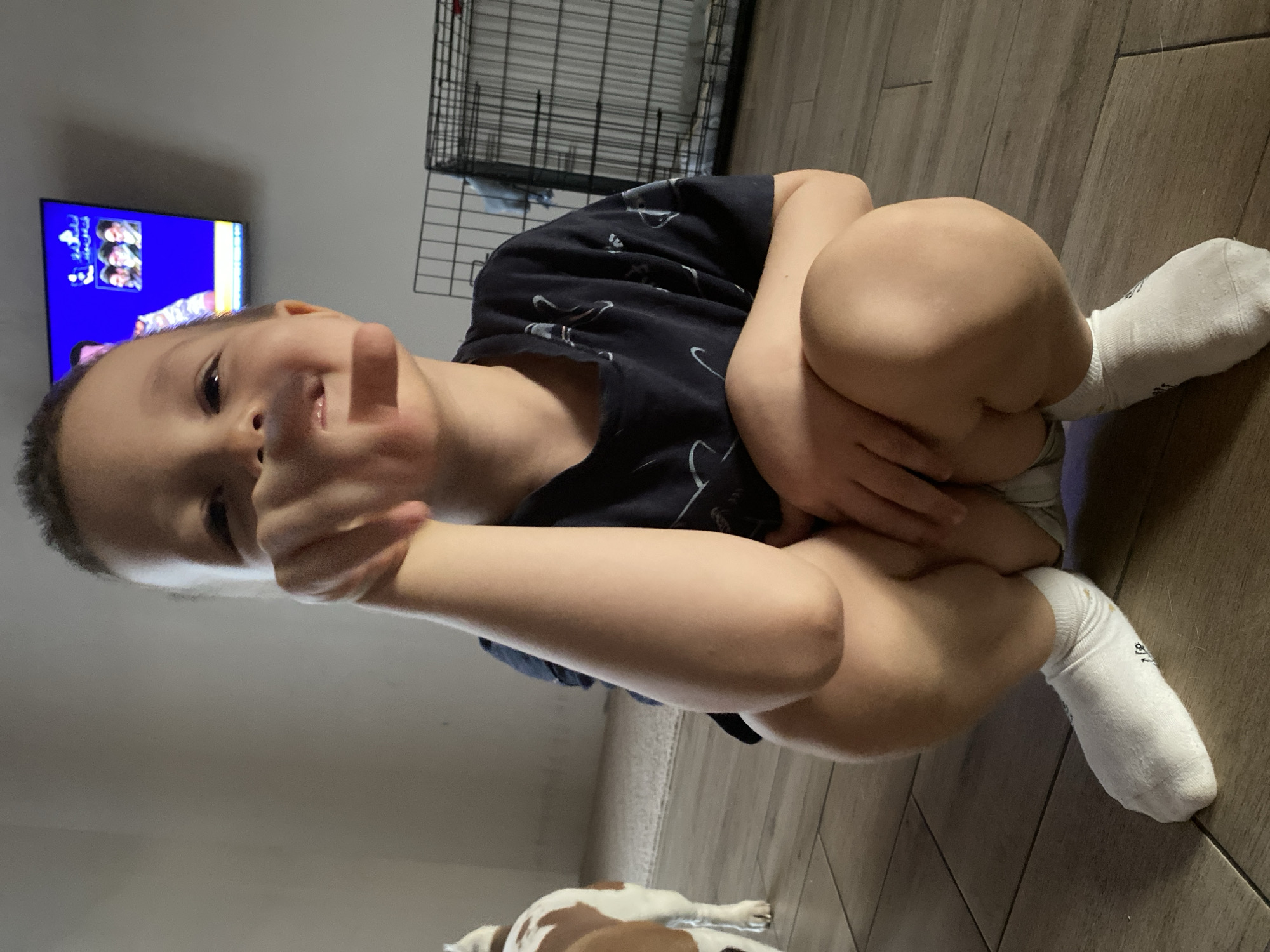 A young boy sits cross-legged on a wooden floor. He wears a dark t-shirt and white socks. He is giving a thumbs-up gesture towards the camera. A television is visible in the background, displaying a colorful program. A wire dog crate stands behind the boy. Part of a dog with brown and white fur is visible in the lower part of the image. The scene appears to be indoors. There is no indication of location in the city of Chaikovsky, Russia.