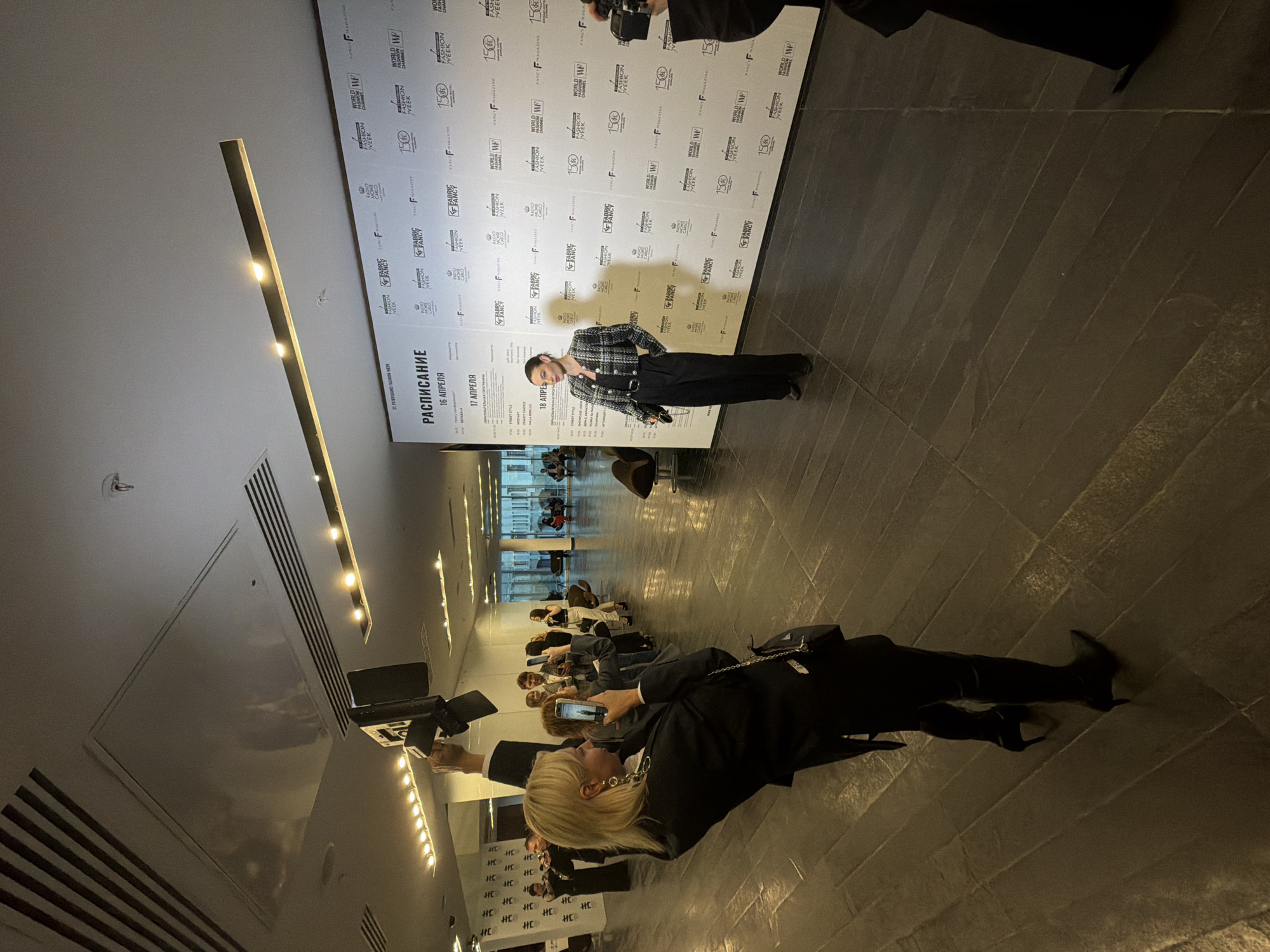 The image captures an indoor event, likely a fashion week or a similar presentation, in what appears to be a modern venue with a dark, reflective tiled floor and a high ceiling. The scene is illuminated by linear ceiling lights and spotlights, one of which is visible in the upper right, held by a person whose upper body is out of frame.
In the foreground, a blonde woman dressed in a black blazer and pants, adorned with a distinctive gold chain earring, is actively recording or photographing the event with her smartphone. Her reflection is clearly visible on the polished floor. Further into the frame, a person in a stylish black and white plaid jacket and dark trousers poses directly in front of a large, branded white backdrop. This backdrop is covered with repeating logos and text, including "WORLD FASHION WEEK," "WF," "FASHION CHANNEL," "FABRIC & FANCY," "F MAGAZINE," and the number "150."
To the left of the person posing, a large schedule board in Russian is prominently displayed. It is titled "РАСПИСАНИЕ" (Schedule) and states "SAINT PETERSBURG FASHION WEEK." Dates for "16 АПРЕЛЯ" (April 16), "17 АПРЕЛЯ" (April 17), and "18 АПРЕЛЯ" (April 18) are visible, indicating a multi-day event. In the mid-ground and background, several other attendees are seated on chairs, some also engrossed in their phones, observing the event. The overall atmosphere suggests a professional and engaged audience at a fashion-related gathering in Saint Petersburg, Russia.