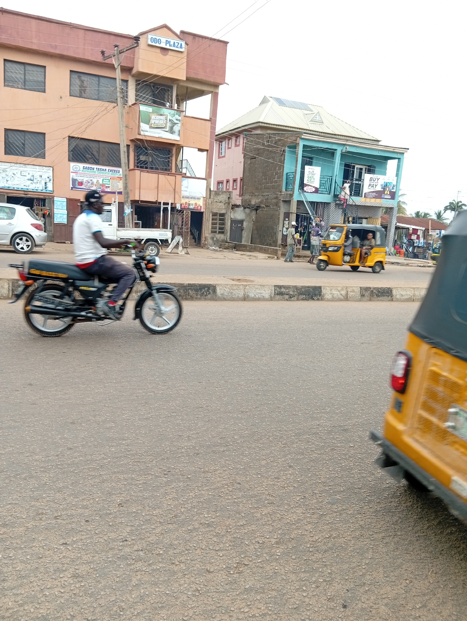 The image shows a street scene in Sabon Tasha, Nigeria. A black motorcycle with a rider wearing a white shirt and a cap is in motion on the left side of the frame. Several buildings are visible in the background, including one with the sign "ODO-PLAZA". Other signs are also present on the buildings. A yellow tricycle (auto-rickshaw) is approaching on the right side of the frame, while another tricycle is further down the street. A few pedestrians are standing near the buildings on the right side of the street. The street is paved.