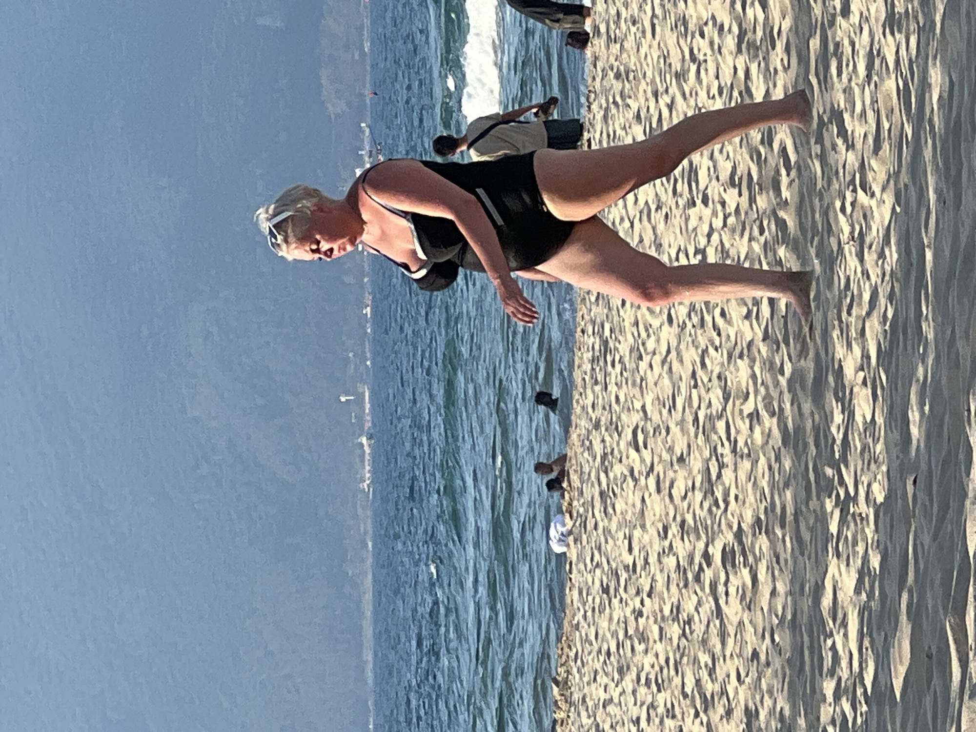 This image depicts a beach scene in Đà Nẵng, Vietnam. The primary subject is a woman in a black swimsuit, standing on the sandy beach. She has blonde hair and is wearing sunglasses. Her mouth is open as if she is speaking or reacting to something. Her body is angled, with her left leg extended. In the background, the ocean with waves is visible, and further out, a hazy coastline with some structures can be seen. Other people are present on the beach and in the water, though they are less prominent. One person, in a light-colored shirt and dark shorts, is walking away from the camera, while another person in darker attire is closer to the water. The sand is textured with shadows, suggesting it is daytime. The water appears calm, with some whitecaps near the shore. The sky is a clear, pale blue. The overall impression is a relaxed day at the beach, with the woman in the foreground being the main focal point of the image. There is no visible text in the image.