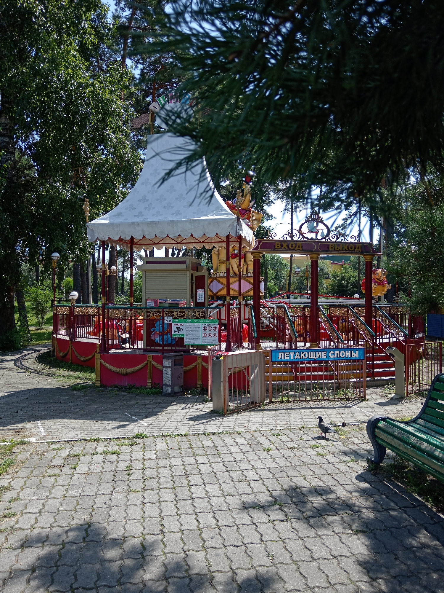 The image depicts a scene in an amusement park, likely located in Belogorsk, Russia, on a sunny day. A carousel with red and gold accents and a white conical roof is the central focus. The carousel appears to be closed, with a shuttered ticket booth visible. A sign above an entrance reads "Вход Выход", meaning "Entrance Exit". Another sign in blue text below the entrance stairs reads "ЛЕТАЮЩИЕ СЛОНЫ", translating to "Flying Elephants".
The amusement ride itself features decorative golden trim and red railings. Several colorful figures, possibly designed to represent animals or mythical creatures, are attached to the carousel's structure. The ride is surrounded by a metal fence.
The environment around the carousel is a park setting, with tall trees providing shade. The ground is paved with interlocking concrete blocks. In the foreground, a green park bench is visible to the right, and two pigeons are present on the pavement. The overall atmosphere is that of a quiet, operational amusement area on a clear day.