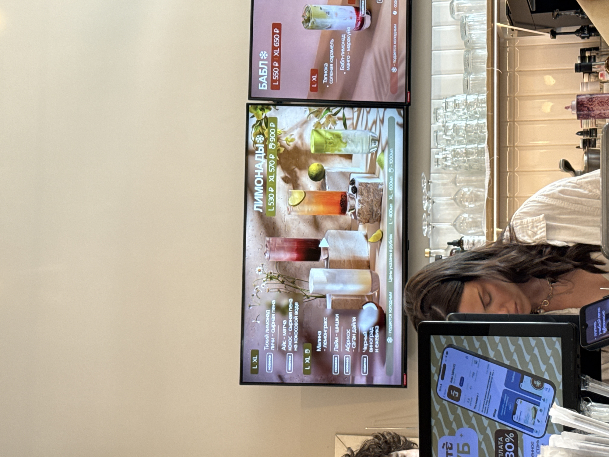 The image shows a cafe interior in Moscow, Russia. In the foreground, a woman with long dark hair is partially visible from the chest up, wearing a white shirt and a necklace. She is looking down, seemingly engaged in her work. To the left of the woman, a computer monitor displays a mobile app interface with Russian text. Above the monitor, a digital menu board is visible, showcasing various drinks with their prices and sizes in Russian. One section of the menu features "ЛИМОНАДЫ" (Lemonades) and another features "БАБЛ" (Bubble Tea). Behind the woman and the counter, shelves are stocked with glassware, and various bottles and containers are present on the counter. The overall environment appears to be a modern cafe setting.