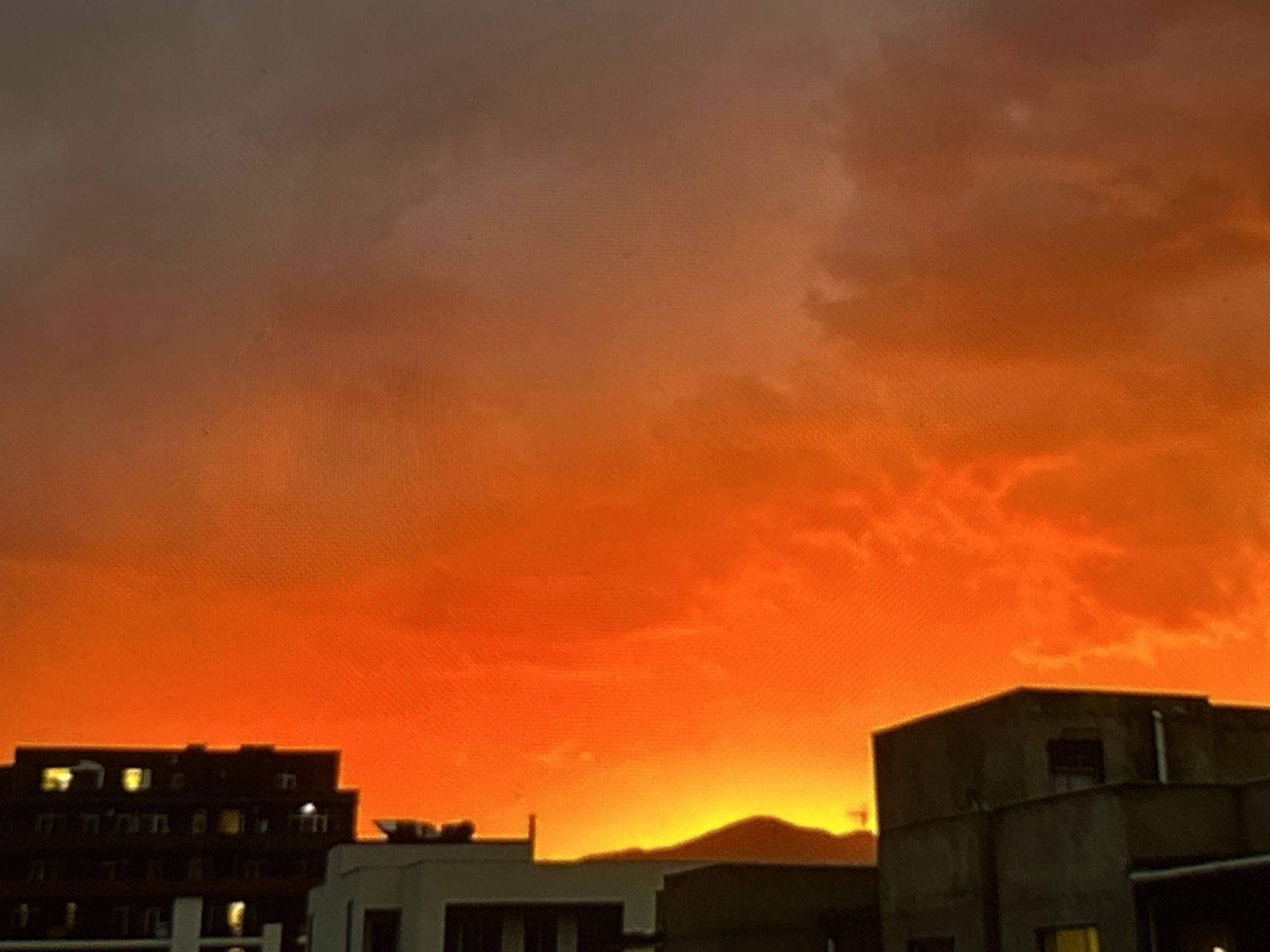 An eye-level shot captures an orange sky over Tbilisi, Georgia. In the foreground, several buildings are silhouetted against the sky. A taller, dark-colored building on the left features multiple windows, some illuminated. To the right, a lighter-colored, smaller building stands beside it. On the far right is a larger, more modern building. The sky is predominantly orange, with darker patches suggesting clouds. In the distance, a mountain is visible against the bright horizon. The overall scene depicts an urban landscape during a sunset or sunrise in Tbilisi, Georgia.